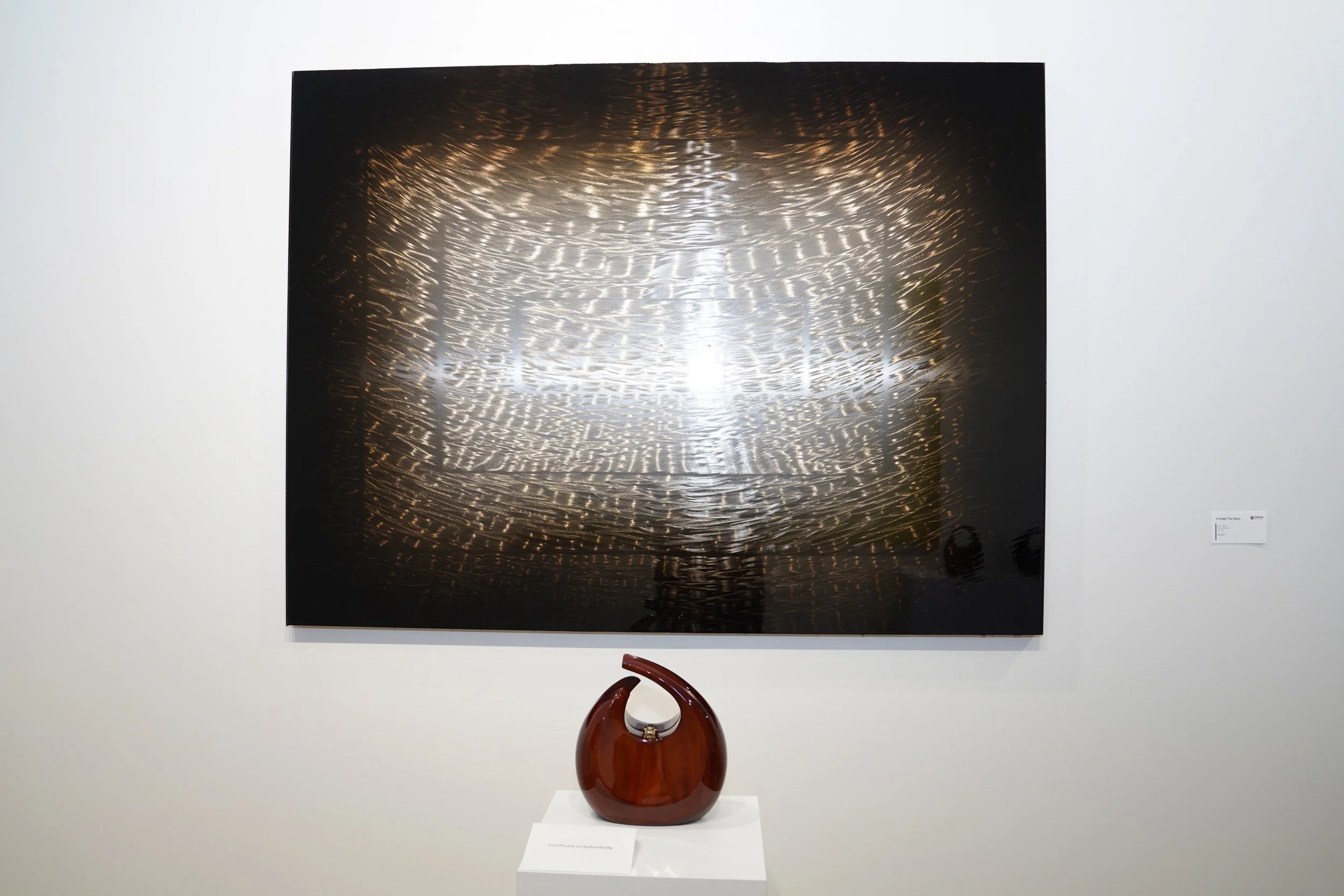 An art gallery featuring a large abstract painting with swirling patterns of metal or reflective material, and a ceramic sculpture on a white pedestal below it.