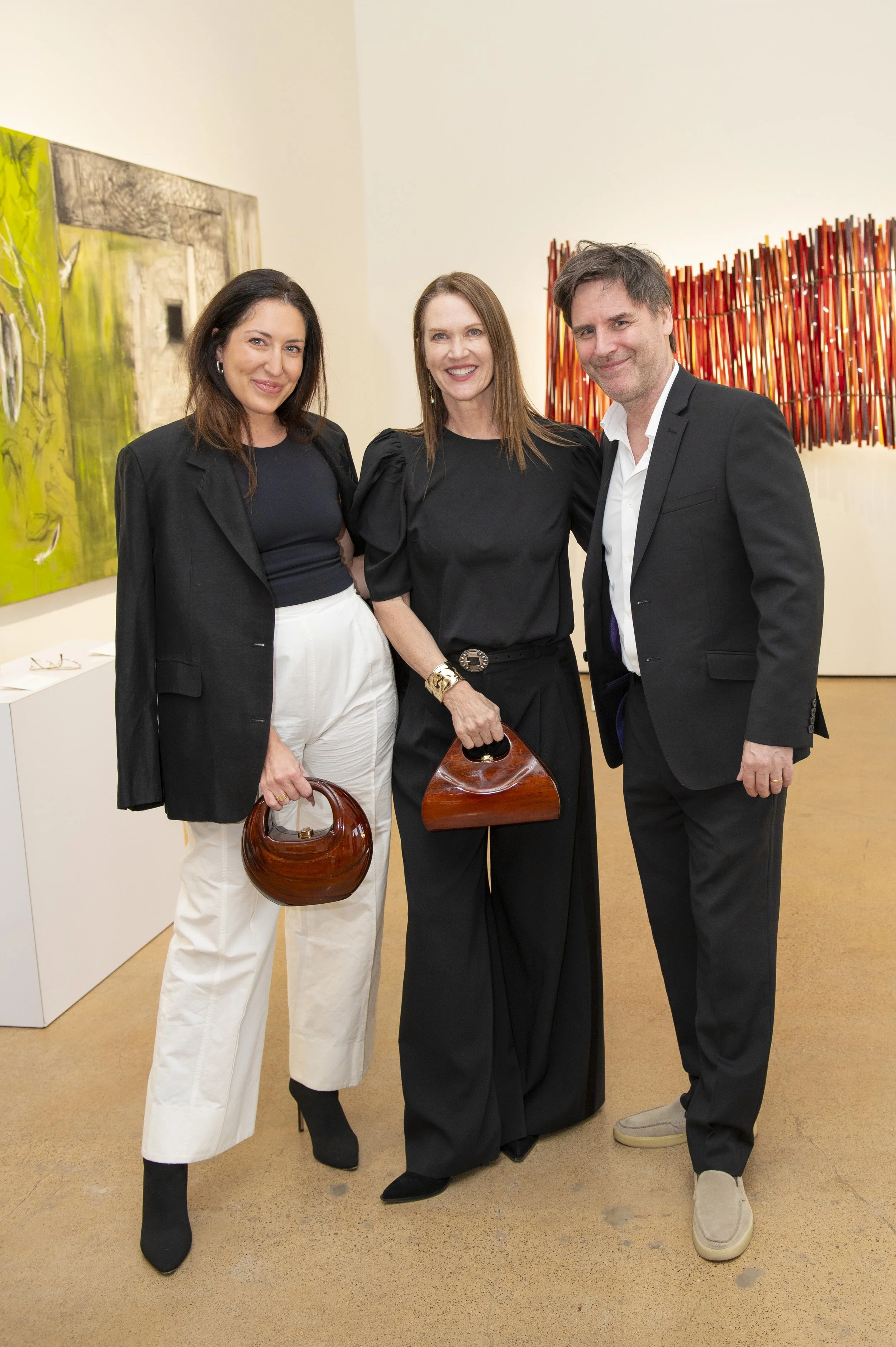 Three people, two women and one man, standing together at an art gallery exhibit, holding wooden handbags, smiling for the camera.