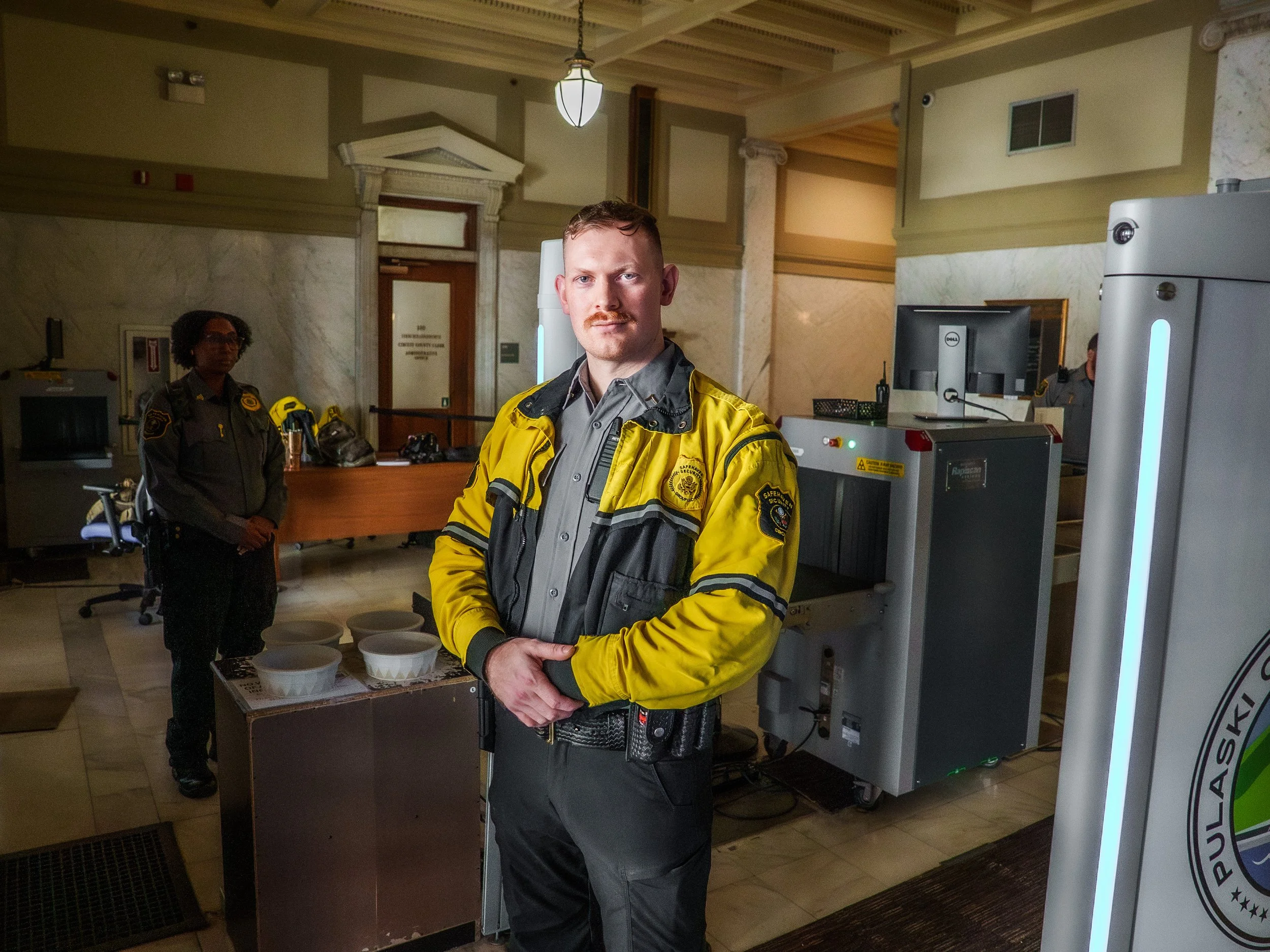 Two uniformed security officers standing in an indoor setting, one in the foreground wearing a yellow jacket and gray shirt, one in the background in darker attire. There are various equipment and boxes around them.