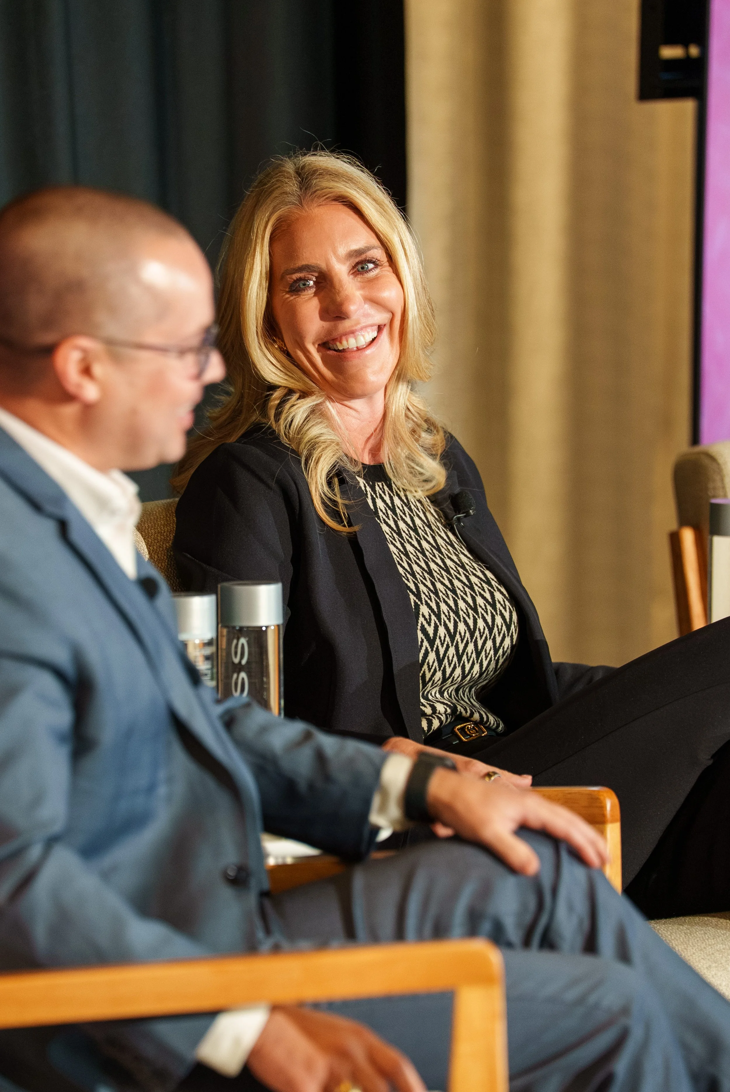 Two people seated at a panel or discussion, a woman smiling and looking to her right, and a man in a suit with glasses looking down, with microphones and water bottles on the table in front of them.