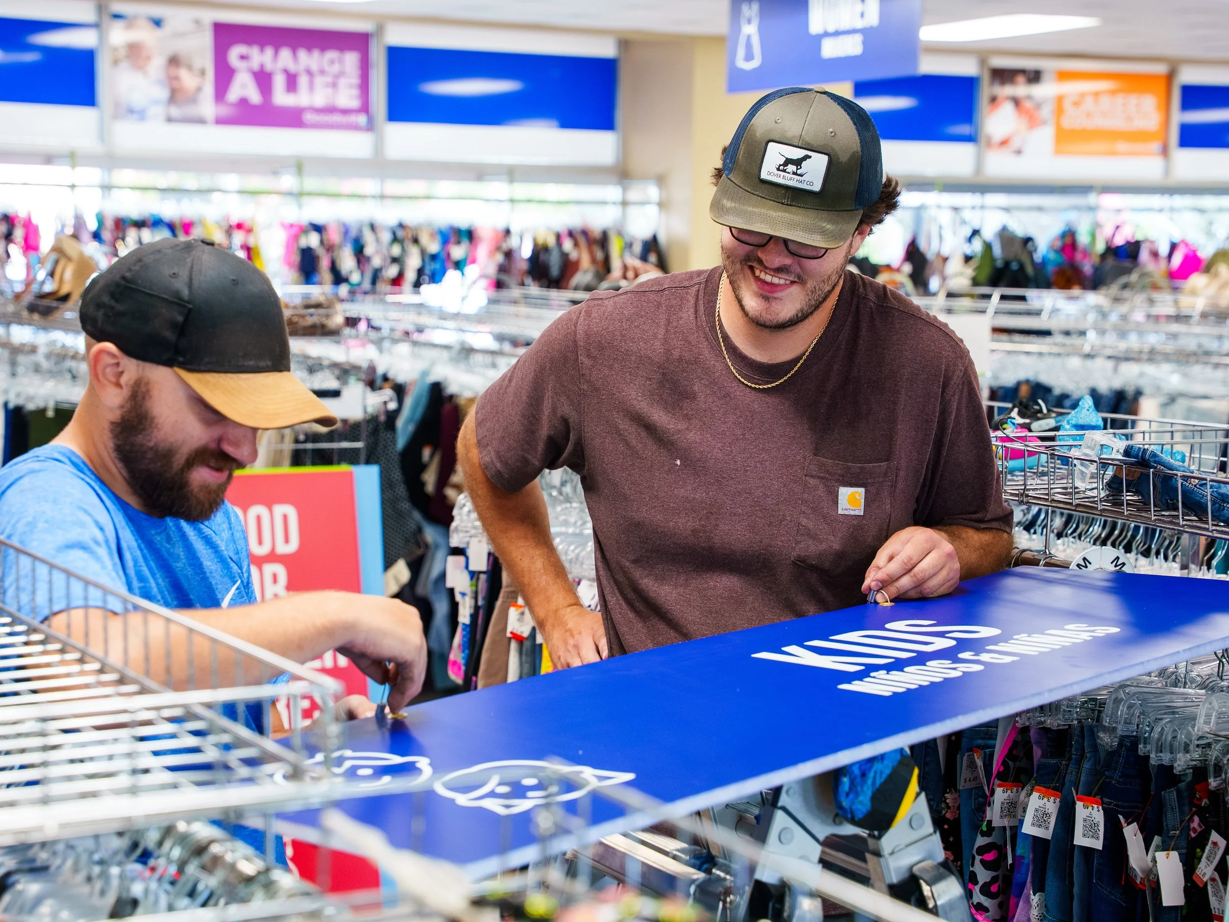 Two men are shopping at a store, one wearing a camouflage cap with a logo and glasses, smiling and looking at items on the counter, while the other wears a black and yellow cap and appears to be shopping with a cart. There are racks of clothing behin