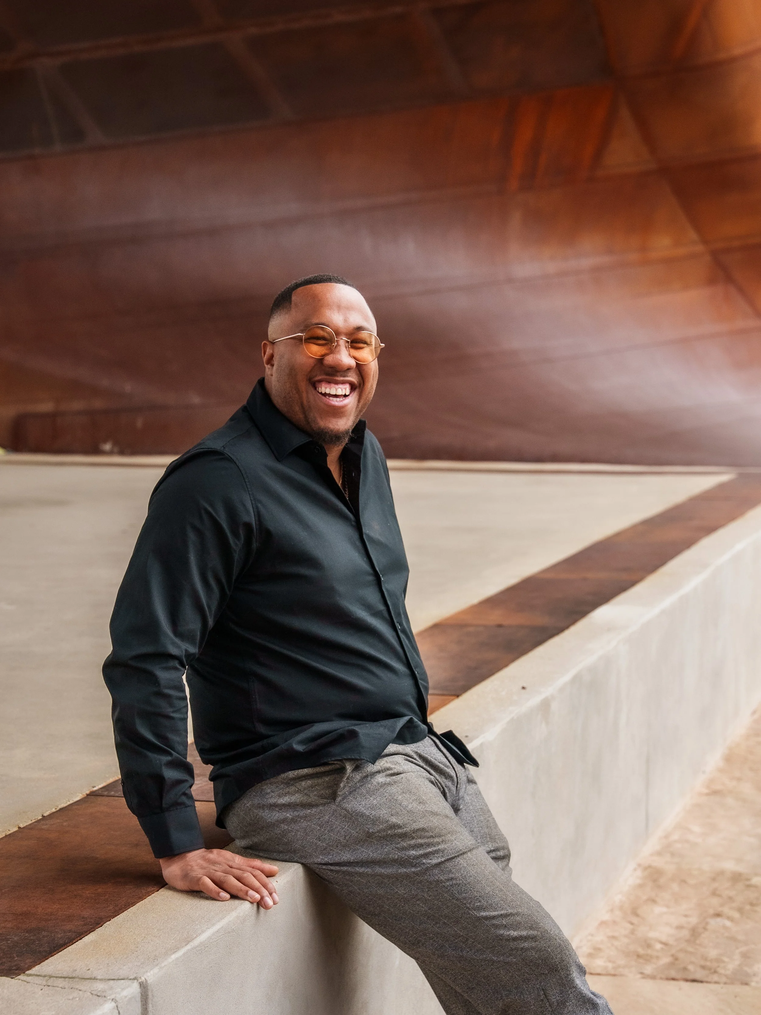 A man with dark skin, wearing glasses with orange-tinted lenses, a black shirt, and gray pants, is smiling and sitting on a curved concrete bench with his right hand resting on it. He is in an indoor setting with a wooden ceiling and beige walls.