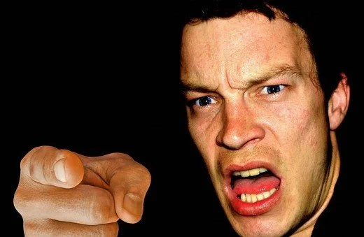 A man point a finger at the camera. His mouth is open and he has an angry look on his face.
