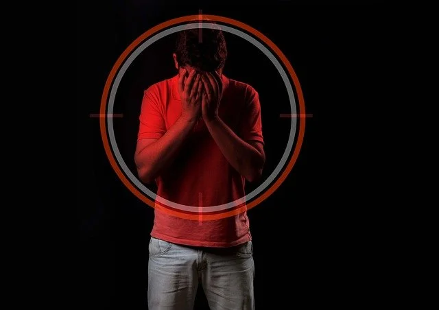 A man against a black background. his head is bowed with his hands in his face. A tageting scope  image is over his trunk and head and has hiscoloured him in a dark red tint.