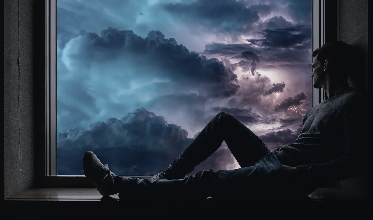 A man sitting in a large windosill in a dark shadow. Looking out the window at stormy clouds.