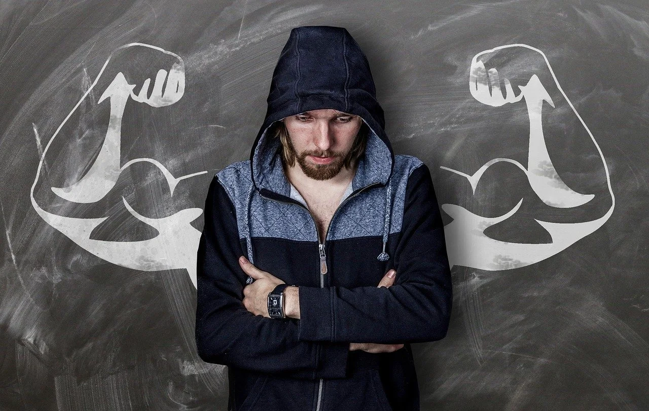 A young man in a hoody arms folded and face turned downward. He is standing in front of a blackboard with a chalk drawing of very muscular arms held up in a weight lifting pose.