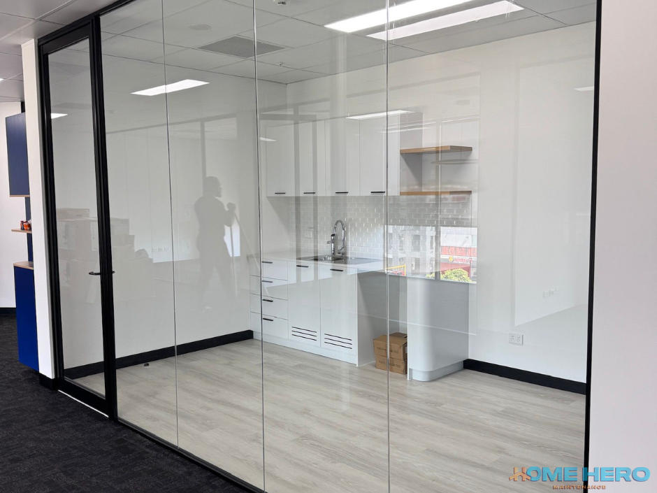 Empty modern kitchen with white cabinets, a sink, and a window, visible through glass walls in an office space.