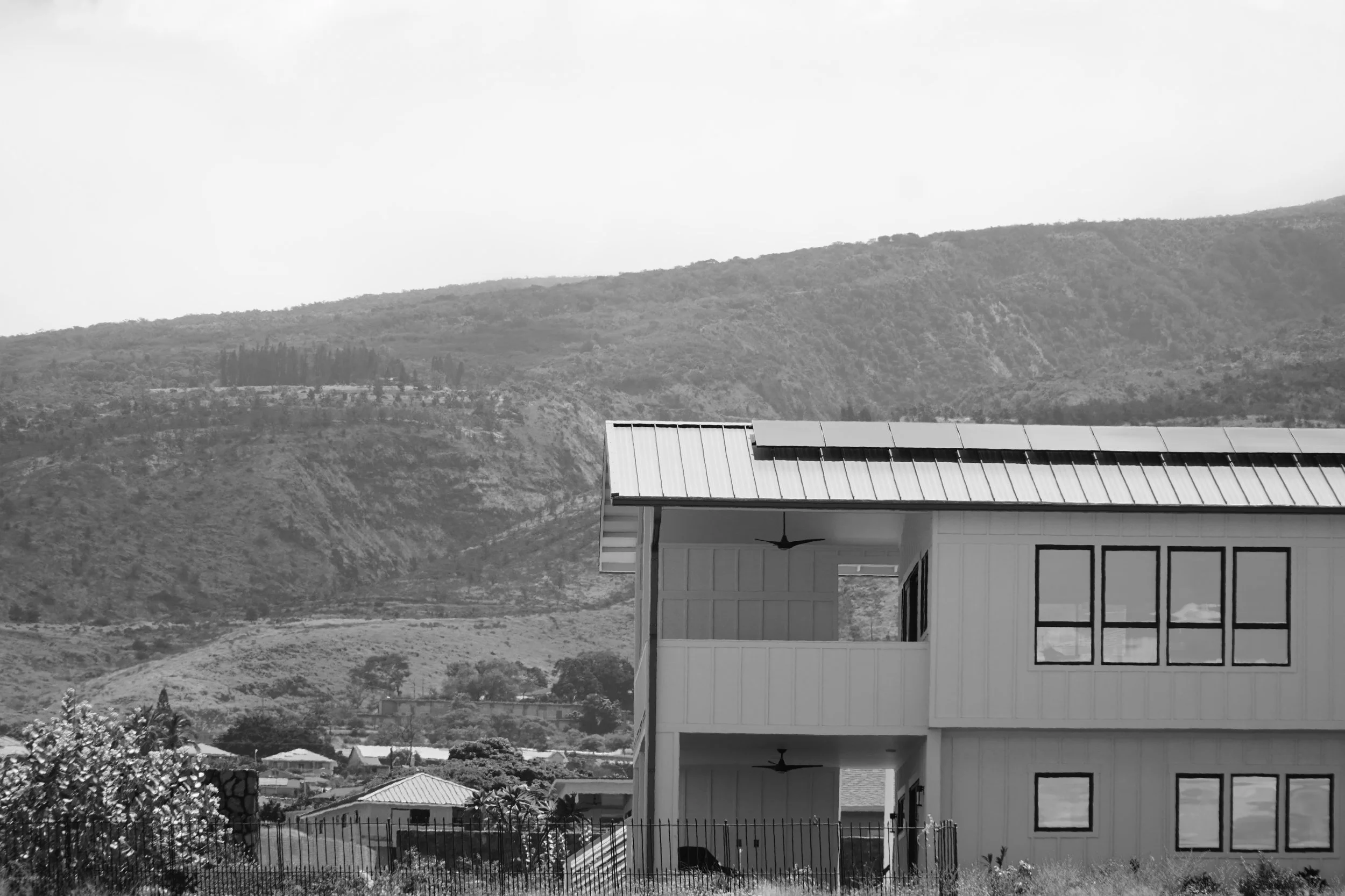 A house with a metal roof and large window panels, situated in a semi-urban area with mountains in the background.