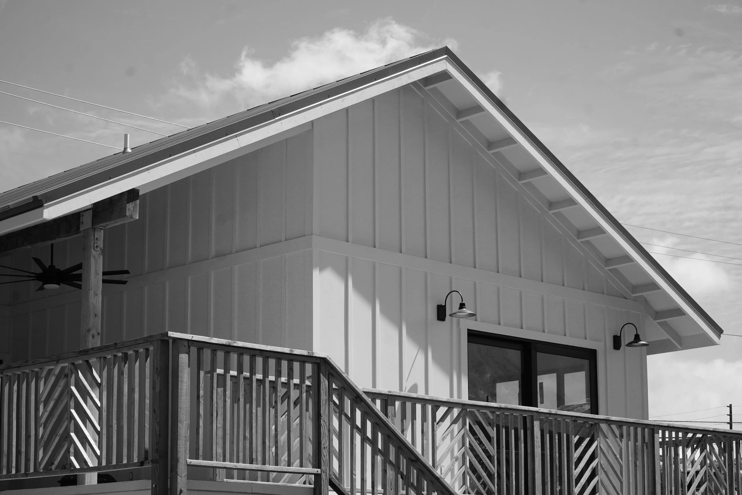 Black and white photo of a house with a steep roof, a railing, and exterior lights, under a partly cloudy sky.