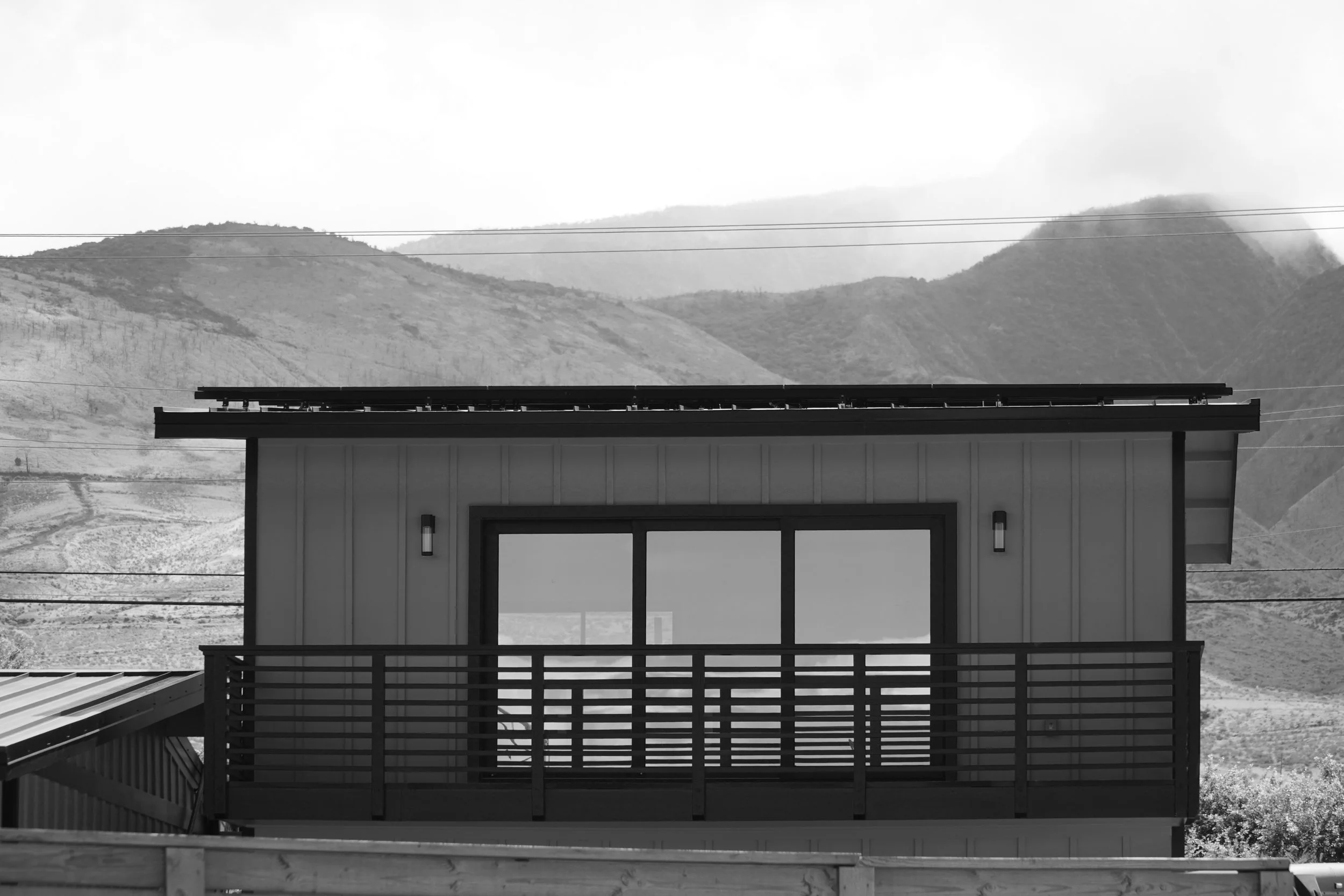 Black and white photo of a modern house with a balcony, set against a mountainous landscape with clouds in the sky.
