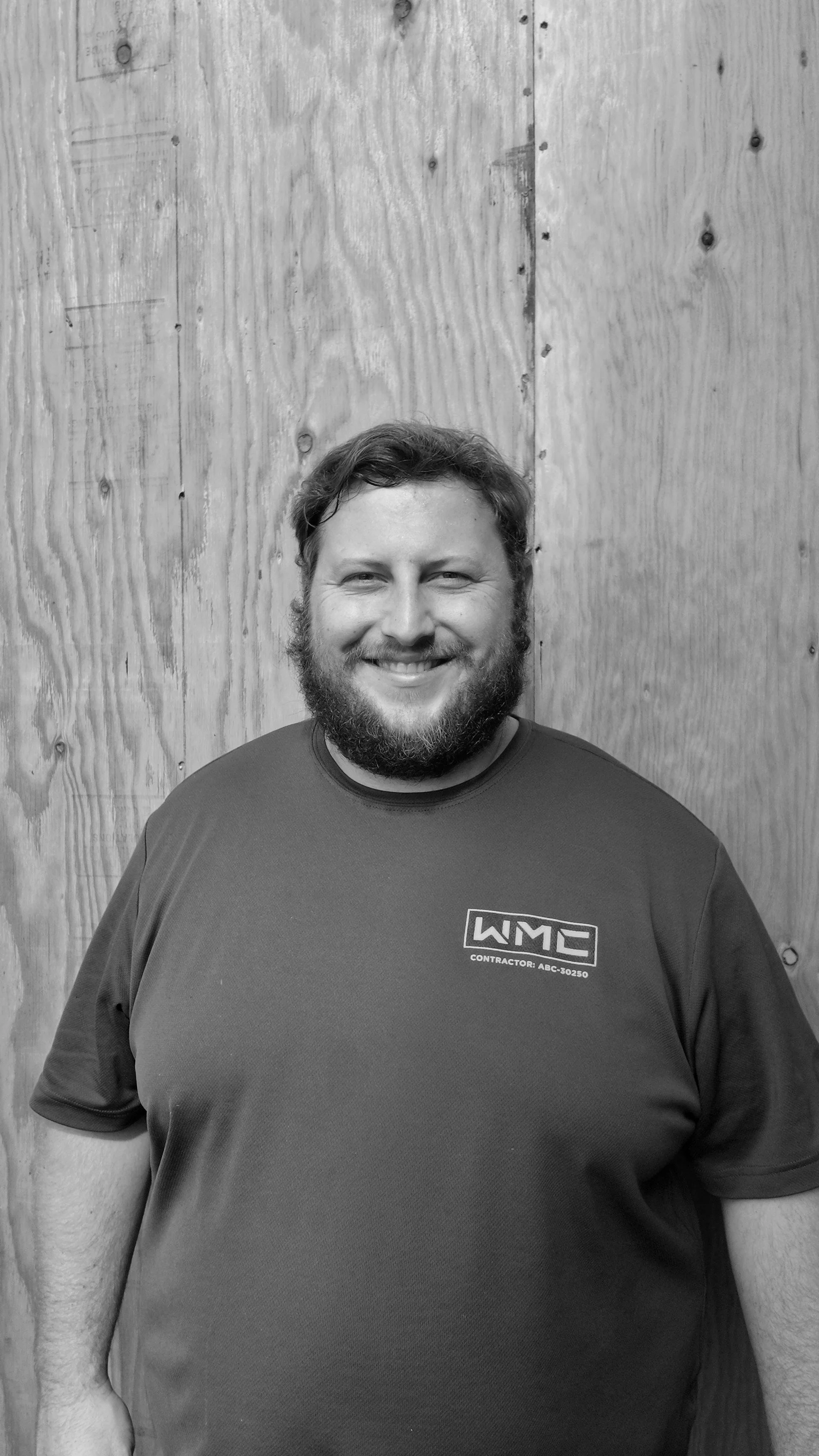 A smiling man with a beard, wearing a dark t-shirt, standing in front of a wooden wall.
