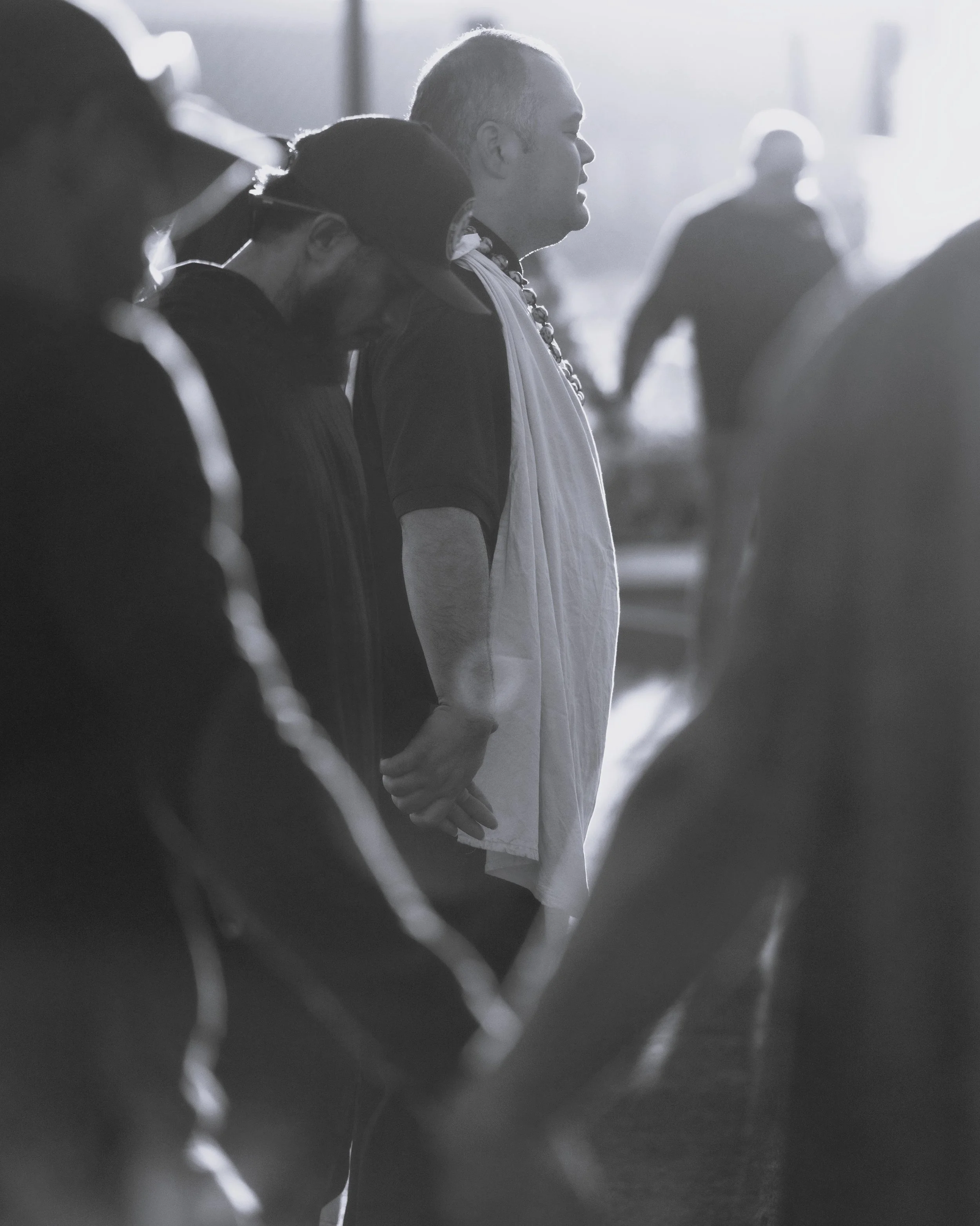 People holding hands in a line, with a man wearing a necklace and a white cloth around his neck, standing outdoors in the sunlight.