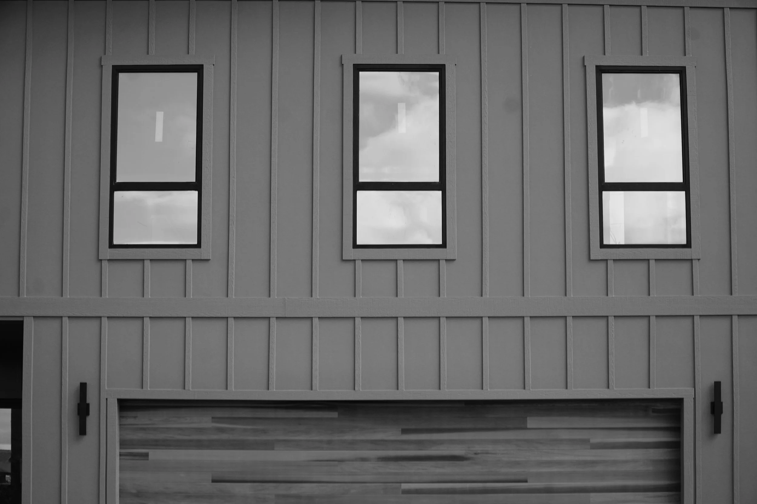 Black and white photo of a building's exterior wall with three vertically aligned windows that reflect a cloudy sky.