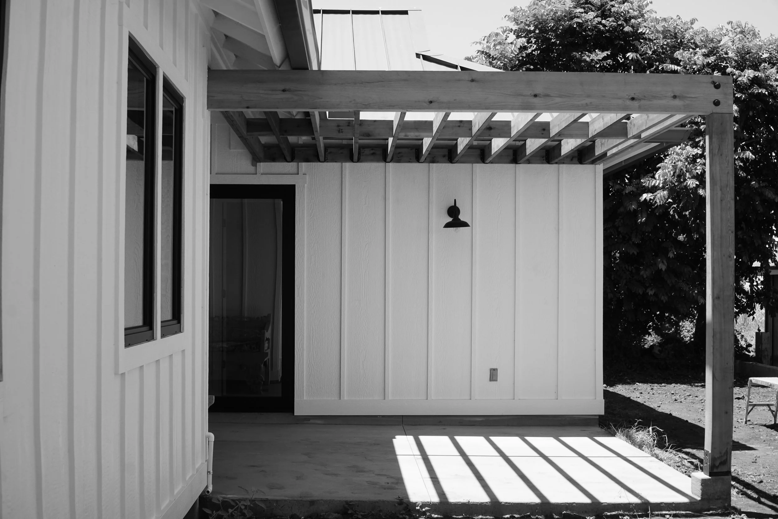 Black and white photo of a house exterior showing a partially completed balcony or deck with wooden framing and vertical siding. A wall-mounted outdoor light is visible beside a glass door, and a tree is in the background.