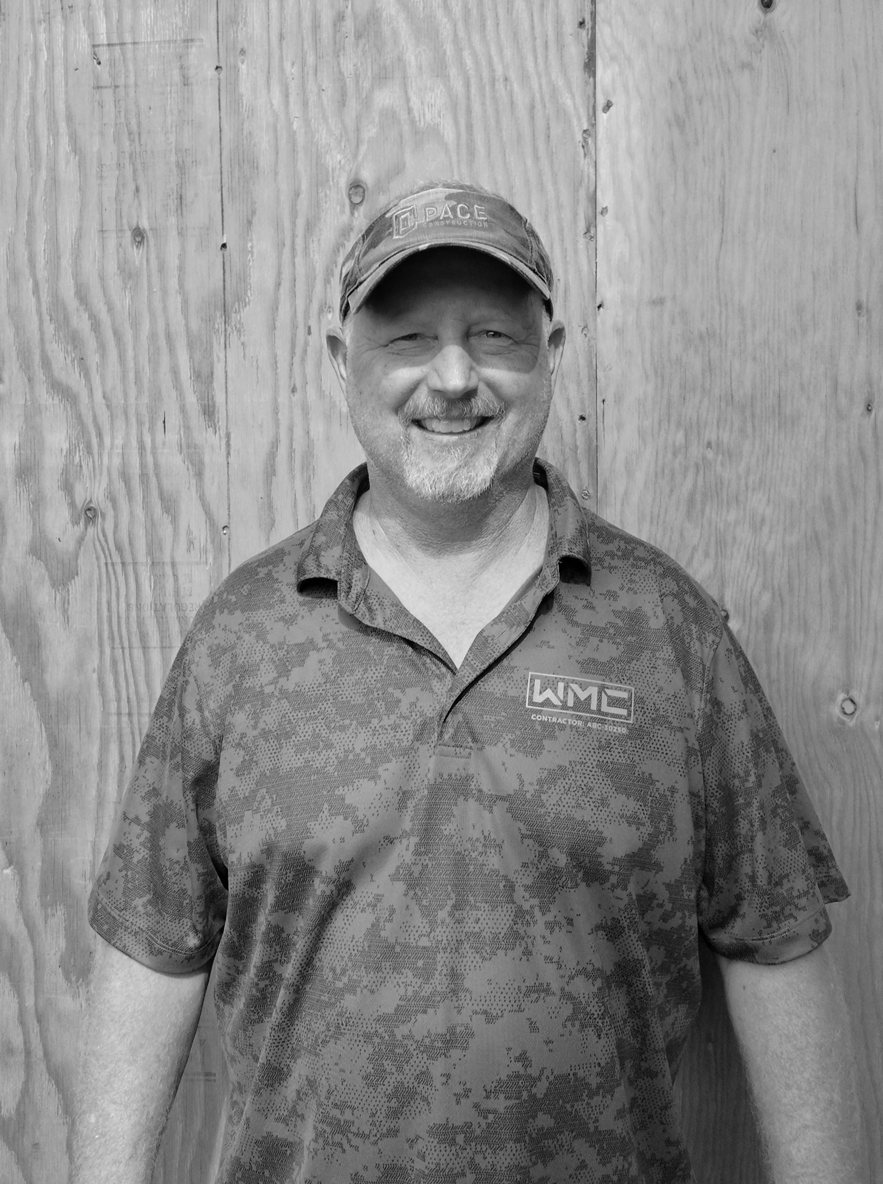 A smiling man wearing a cap and a camouflage polo shirt standing in front of a wooden wall.
