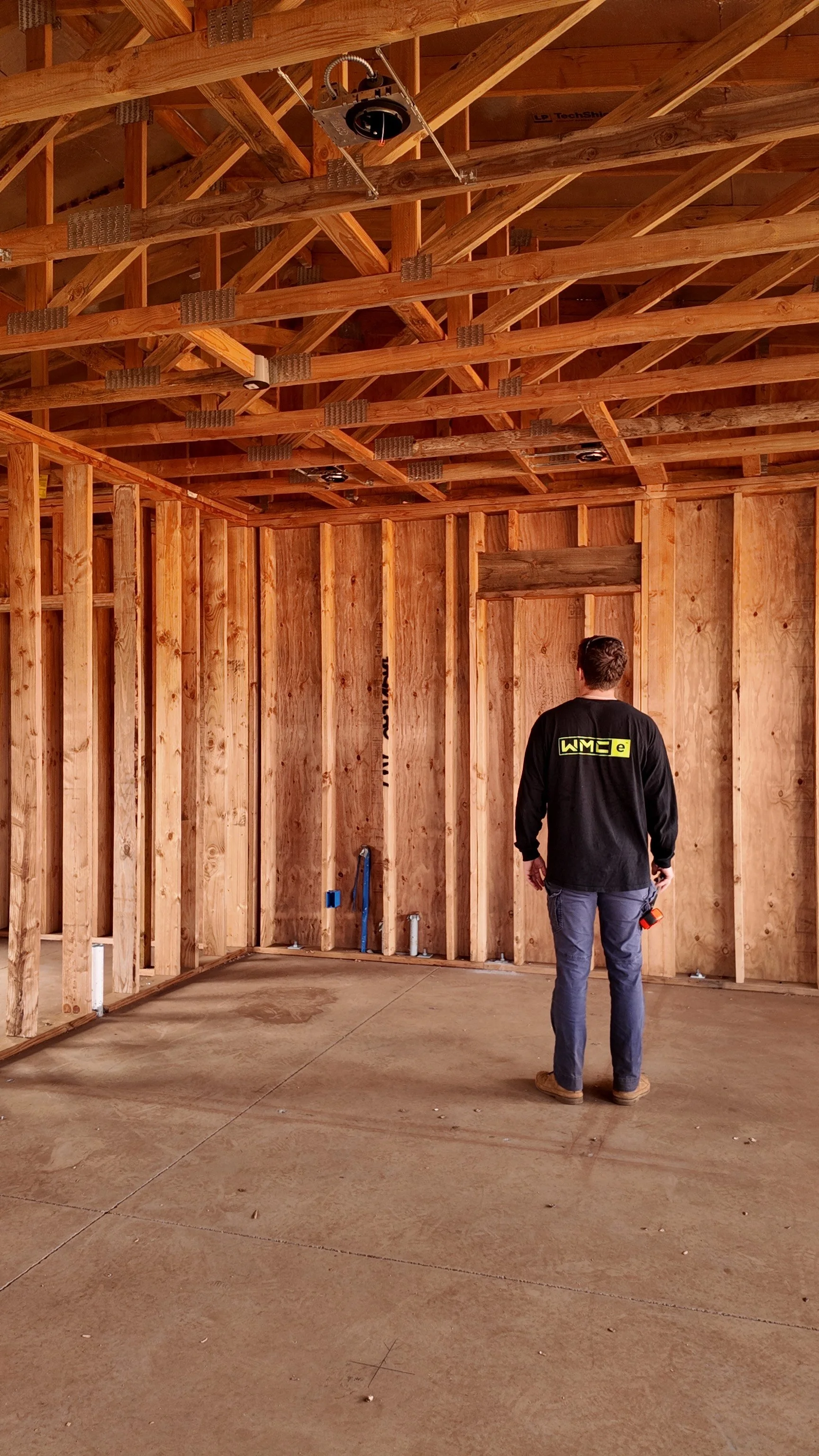 A man inspecting the framed wooden walls and ceiling of an unfinished garage or room under construction, with exposed wood trusses and concrete floor.