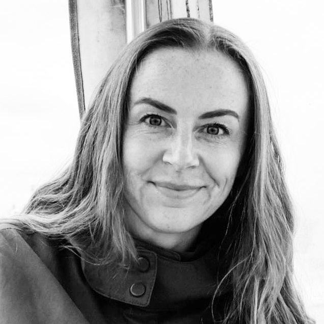 Black and white photo of a woman (the designer Debbie Joy) with long, wavy hair smiling at the camera, wearing a collared jacket.