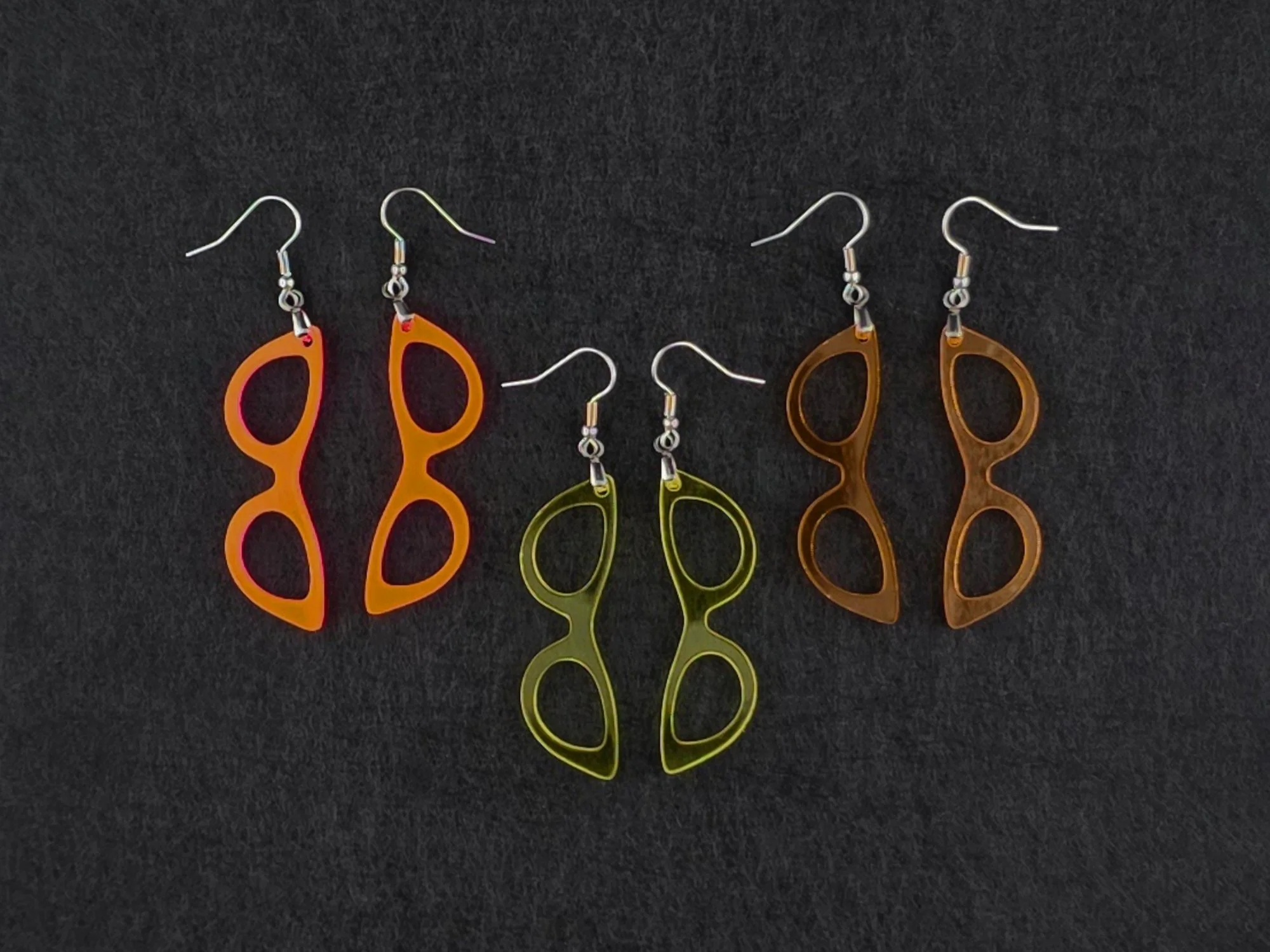 Three pairs of cat-eye earrings on charcoal in fluorescent orange-pink, translucent yellow, and amber orange acrylic, showing three colorways side by side.
