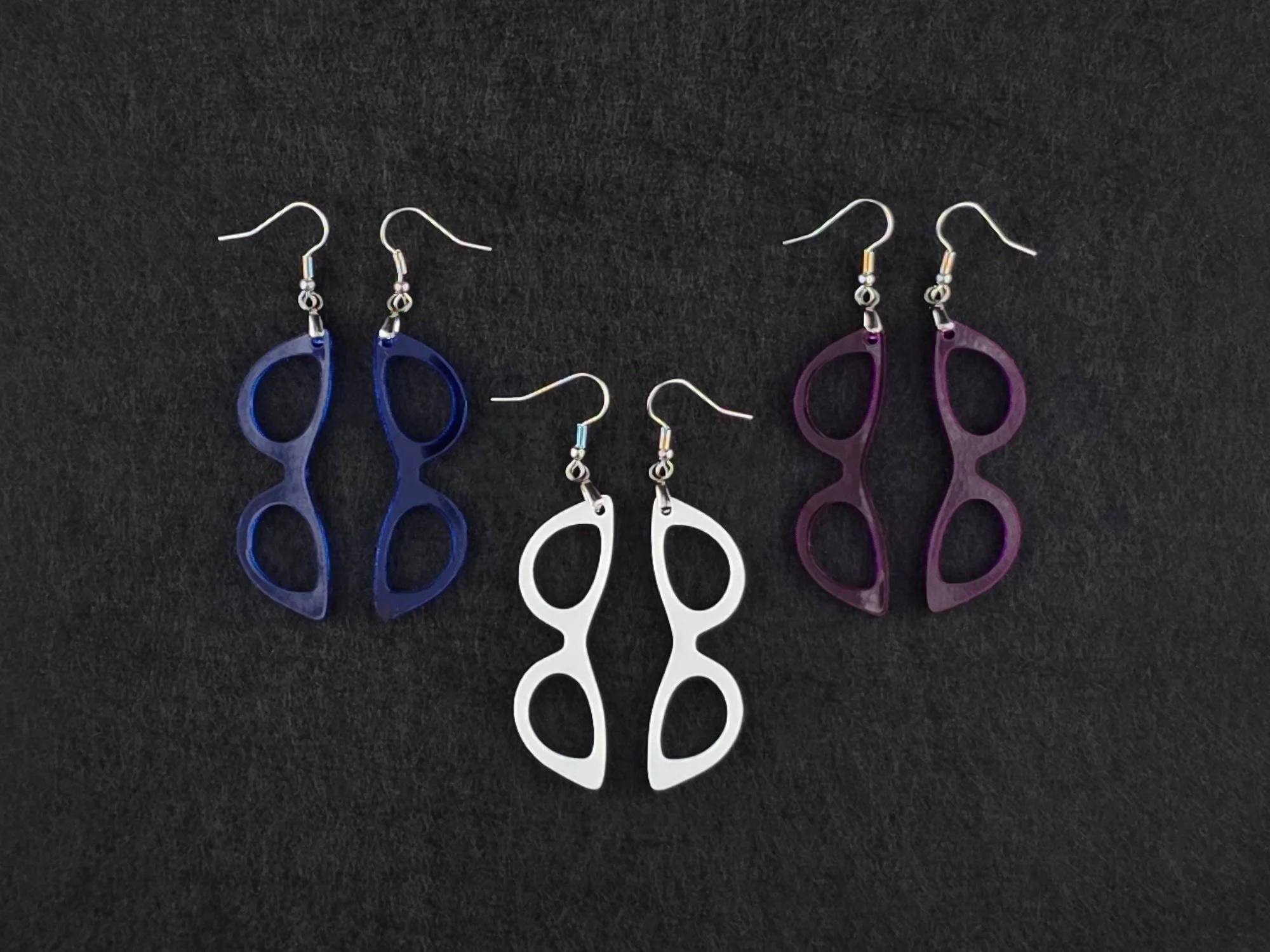Three pairs of cat-eye earrings on charcoal in blue, white, and purple acrylic, showing three more colorways side by side.