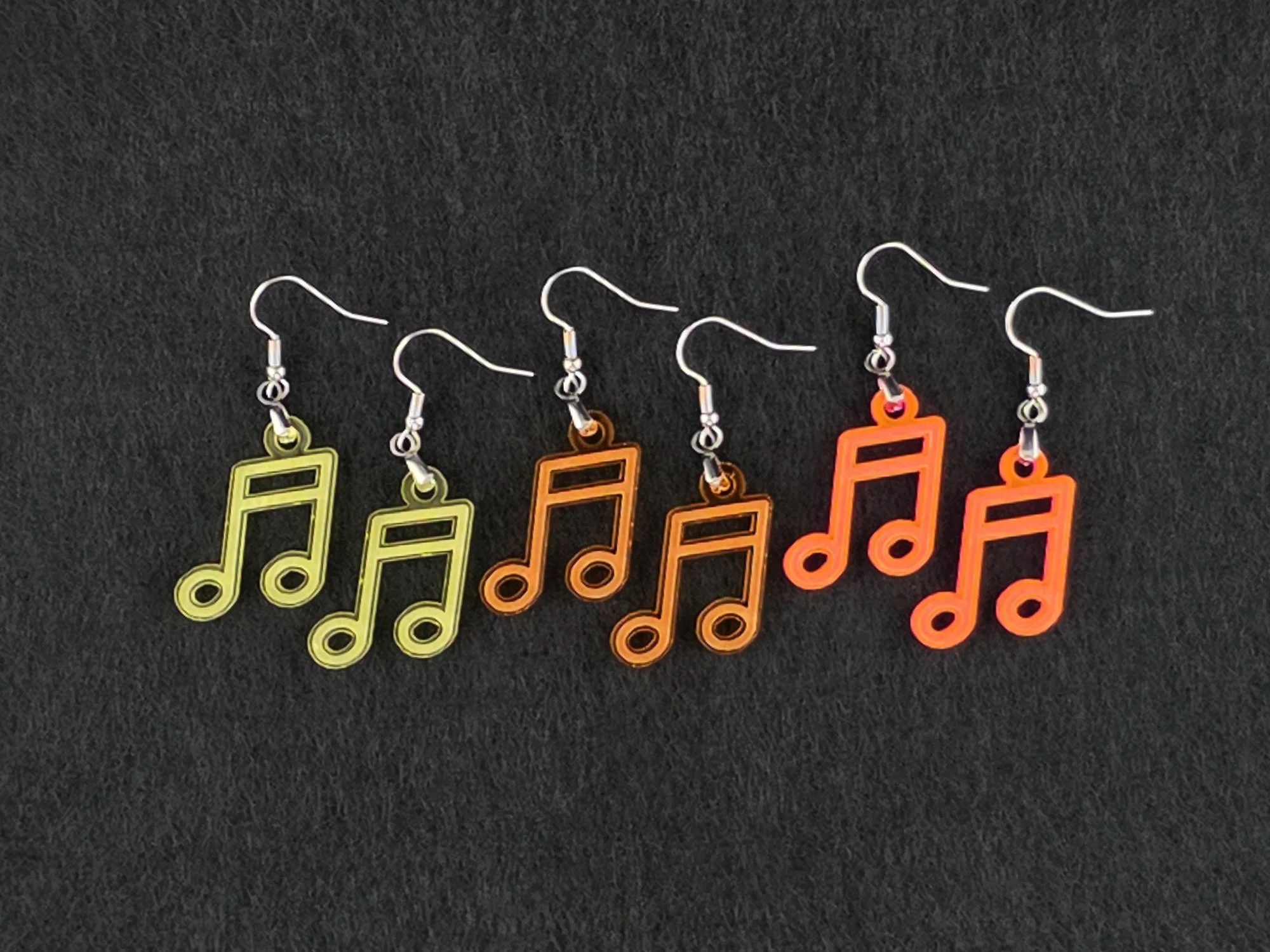 Three pairs of beam note earrings on charcoal in yellow, orange, and fluorescent acrylic, shown side by side for color comparison.