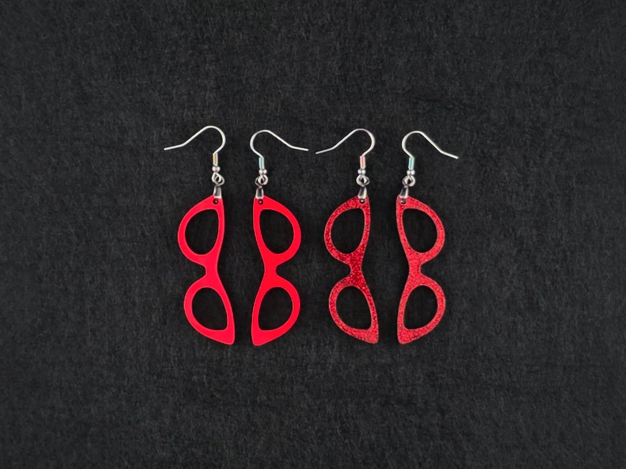 Two pairs of cat-eye earrings side by side on charcoal, one in matte red acrylic and one in a red glitter finish, showing the difference between the two textures.