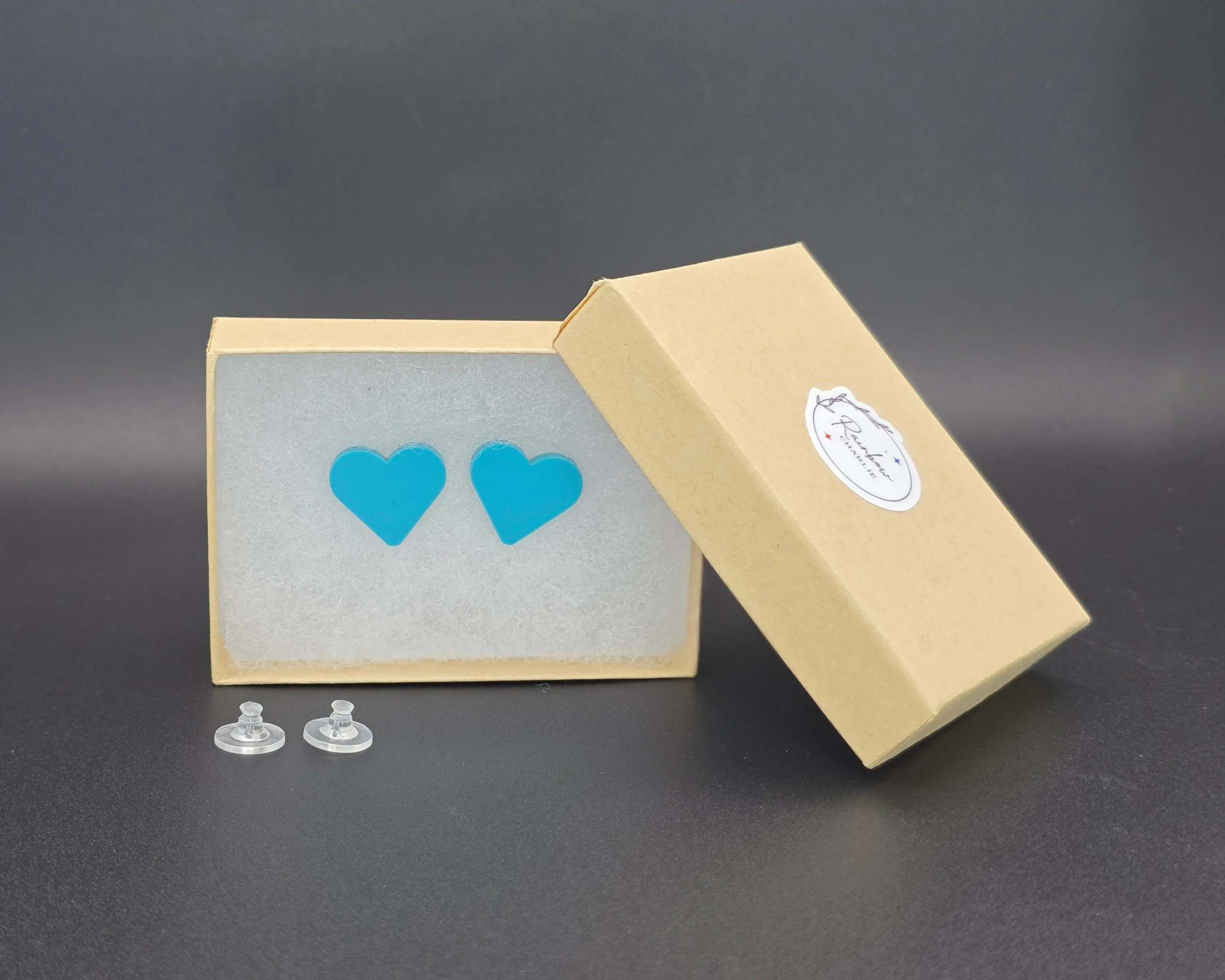 Teal acrylic heart stud earrings packaged in a kraft gift box with Rainbow Charlie branding and silicone earring backs, handmade and ready to gift