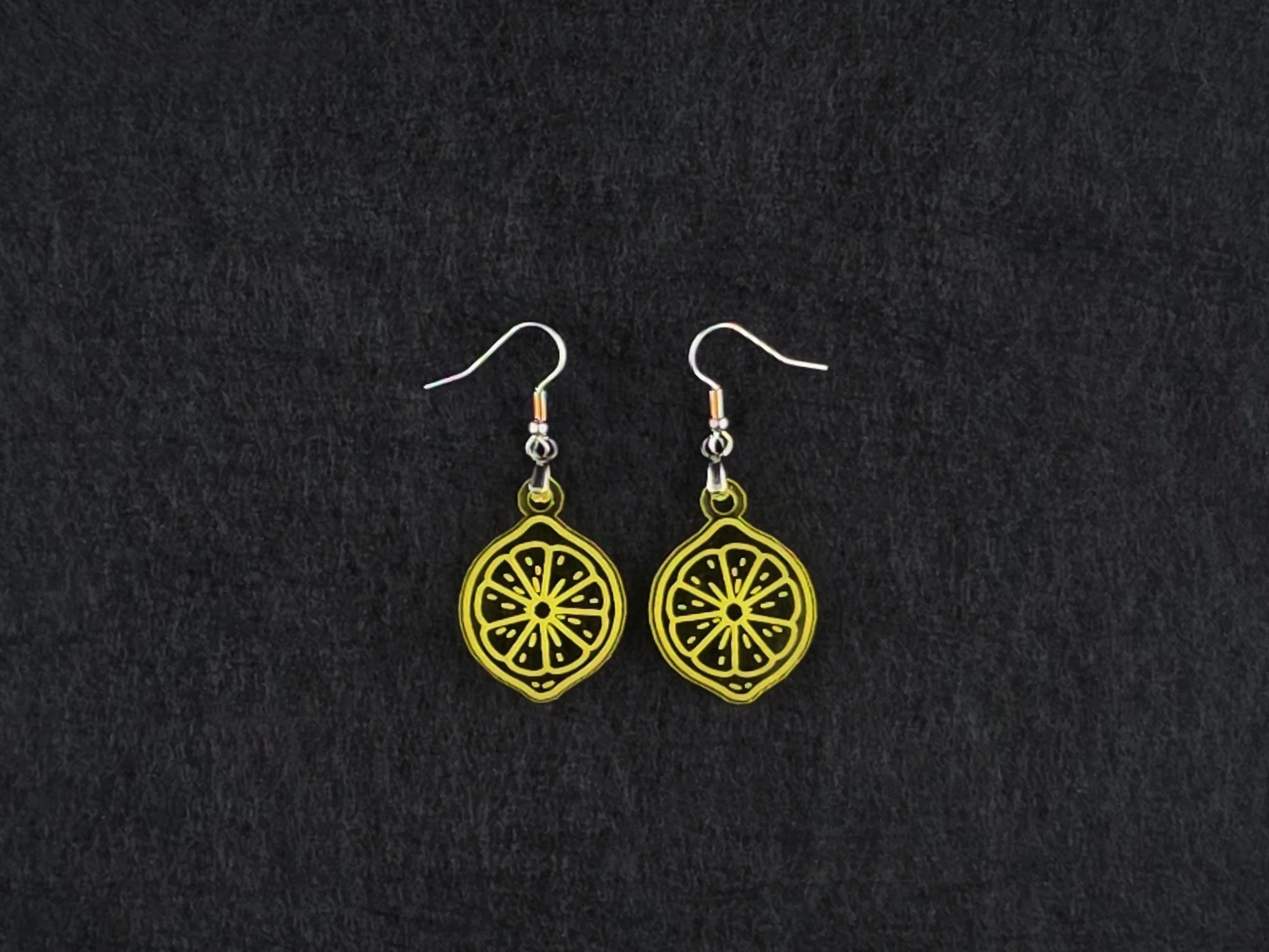 Pair of transparent yellow acrylic lemon slice earrings on stainless steel ear wires, flat lay on a charcoal background.