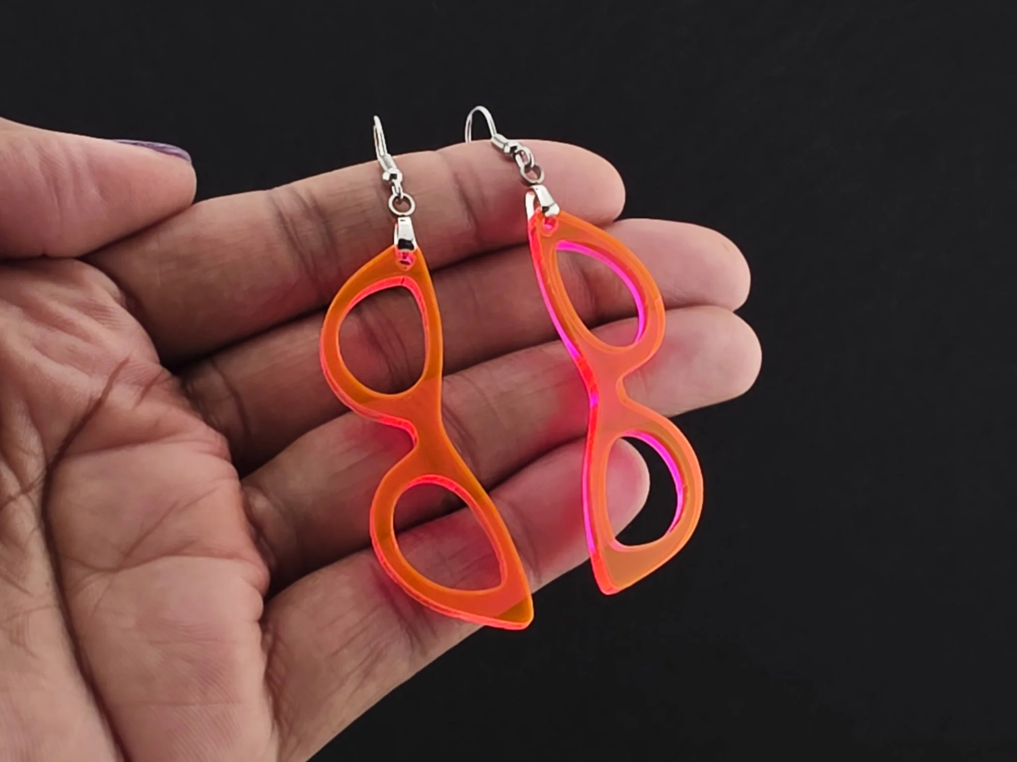 Cat-eye earrings in fluorescent orange acrylic resting in an open palm on a black background, showing the pink glow along the cut edges.