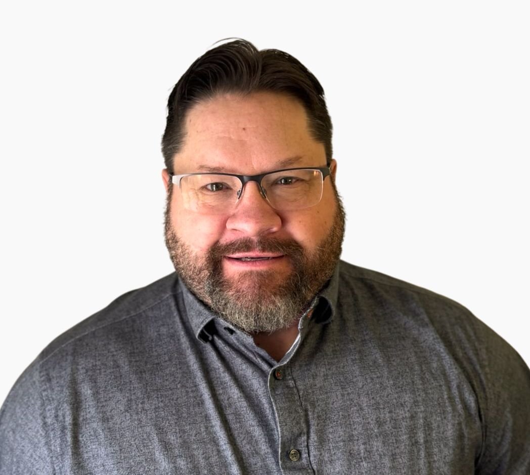 A middle-aged man with glasses and a beard wearing a gray button-up shirt, smiling, against a plain white background.