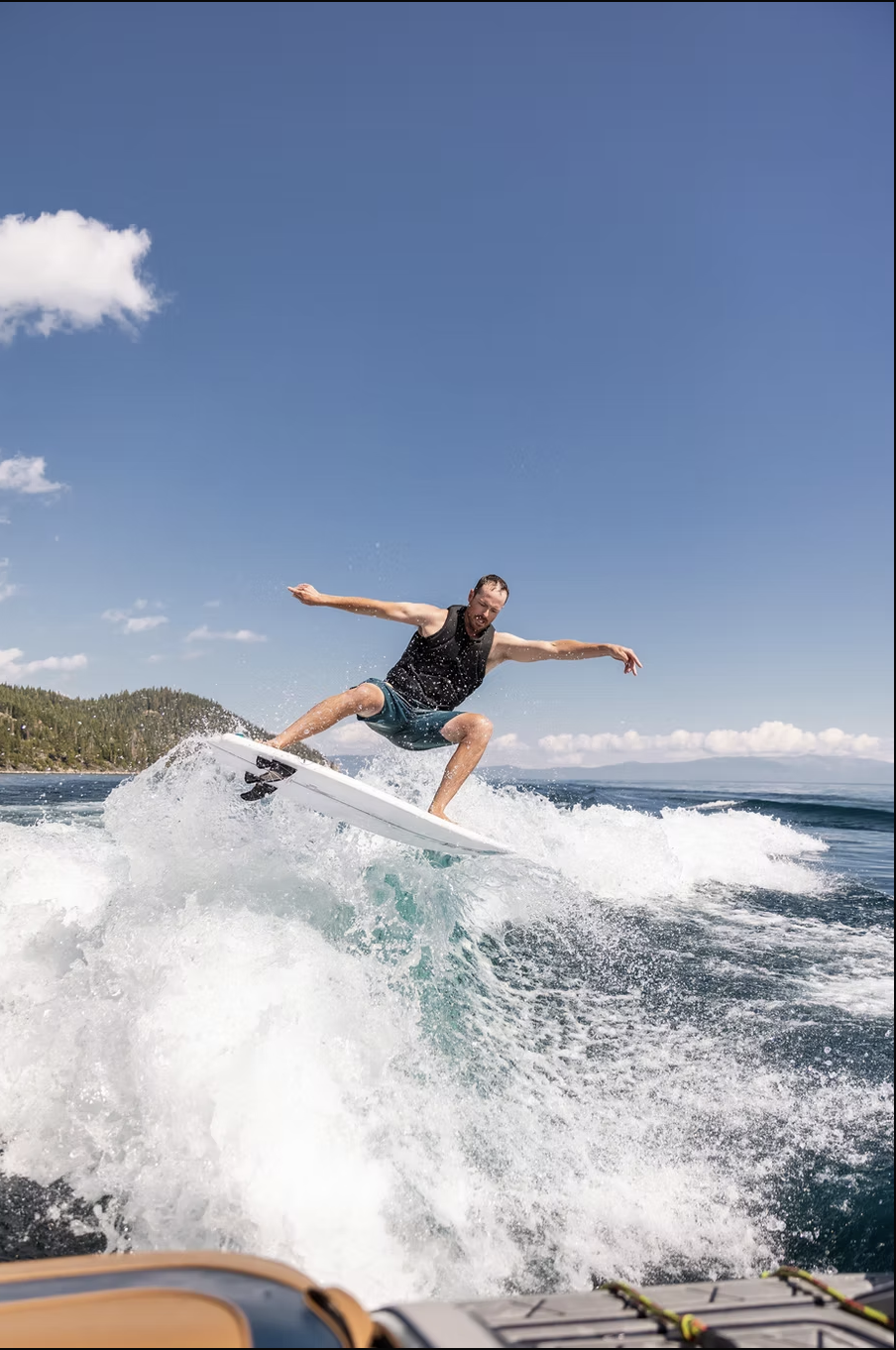 A man wearing a black tank top and blue shorts surfing on a wave in a lake or ocean under a blue sky with some clouds.