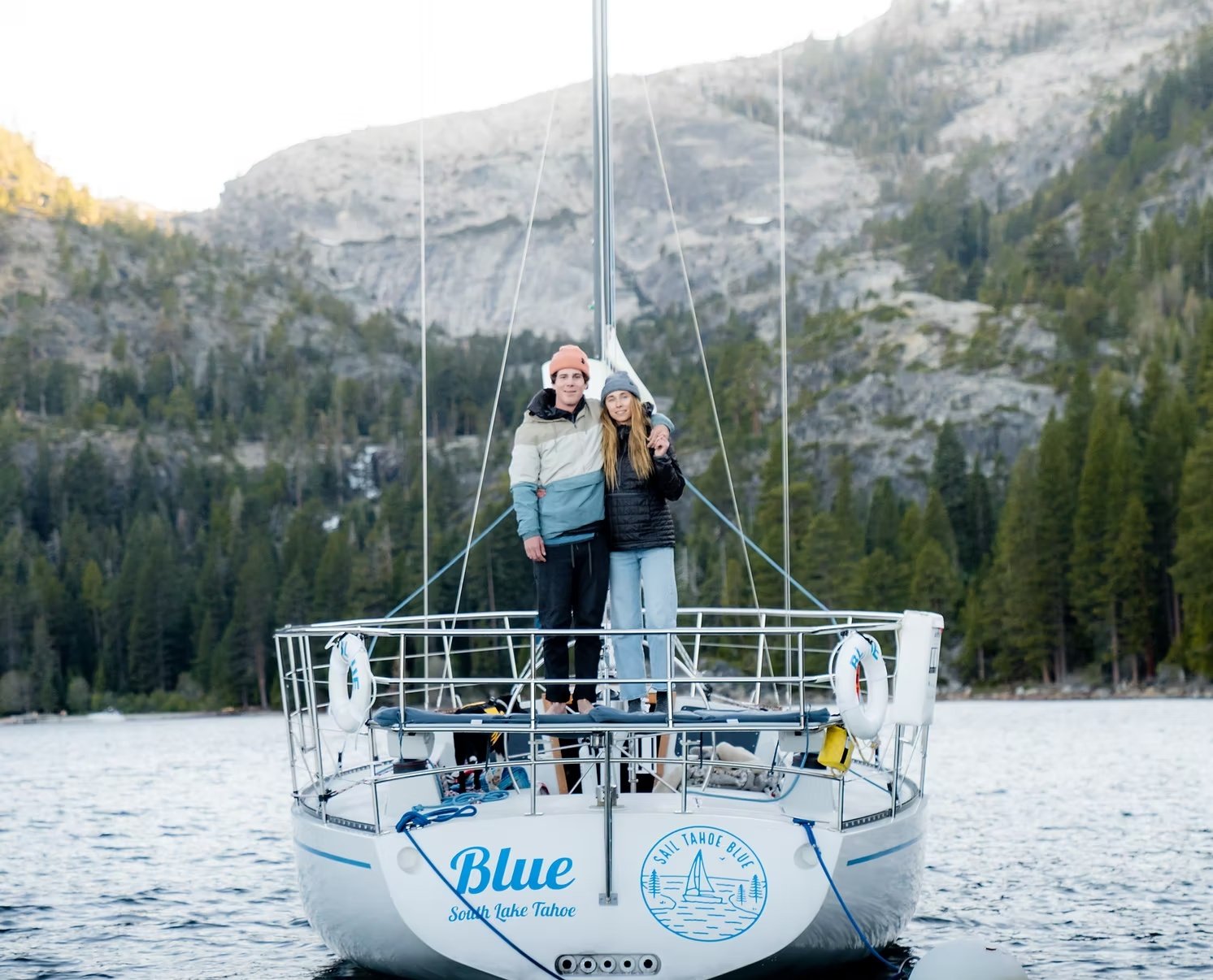 A couple stands on the deck of a sailboat named 'Blue' on South Lake Tahoe, with a mountain and forested shoreline in the background.