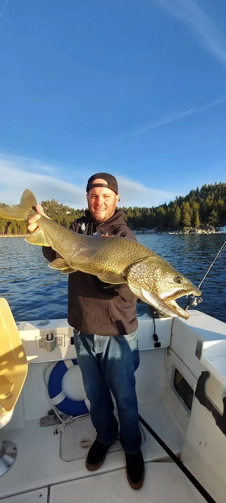 A man standing on a boat holding a large fish, likely a lake trout or similar freshwater species, with a scenic background of a lake, forested shoreline, and a clear blue sky.