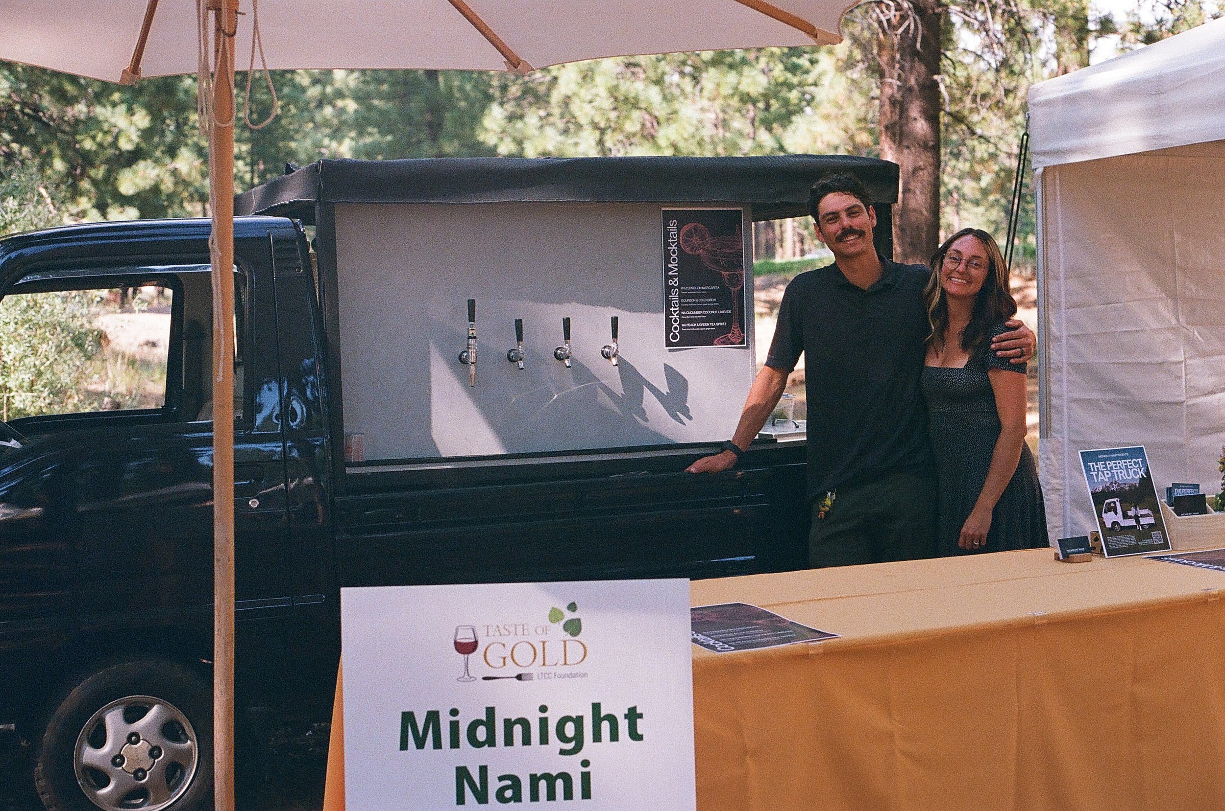 Two people smiling and hugging in front of a drink station at an outdoor event. The station has three taps labeled for cocktails and mocktails. There are trees in the background and part of a white canopy on the right side. A sign on the table reads "Taste of Gold" and "Midnight Nami."