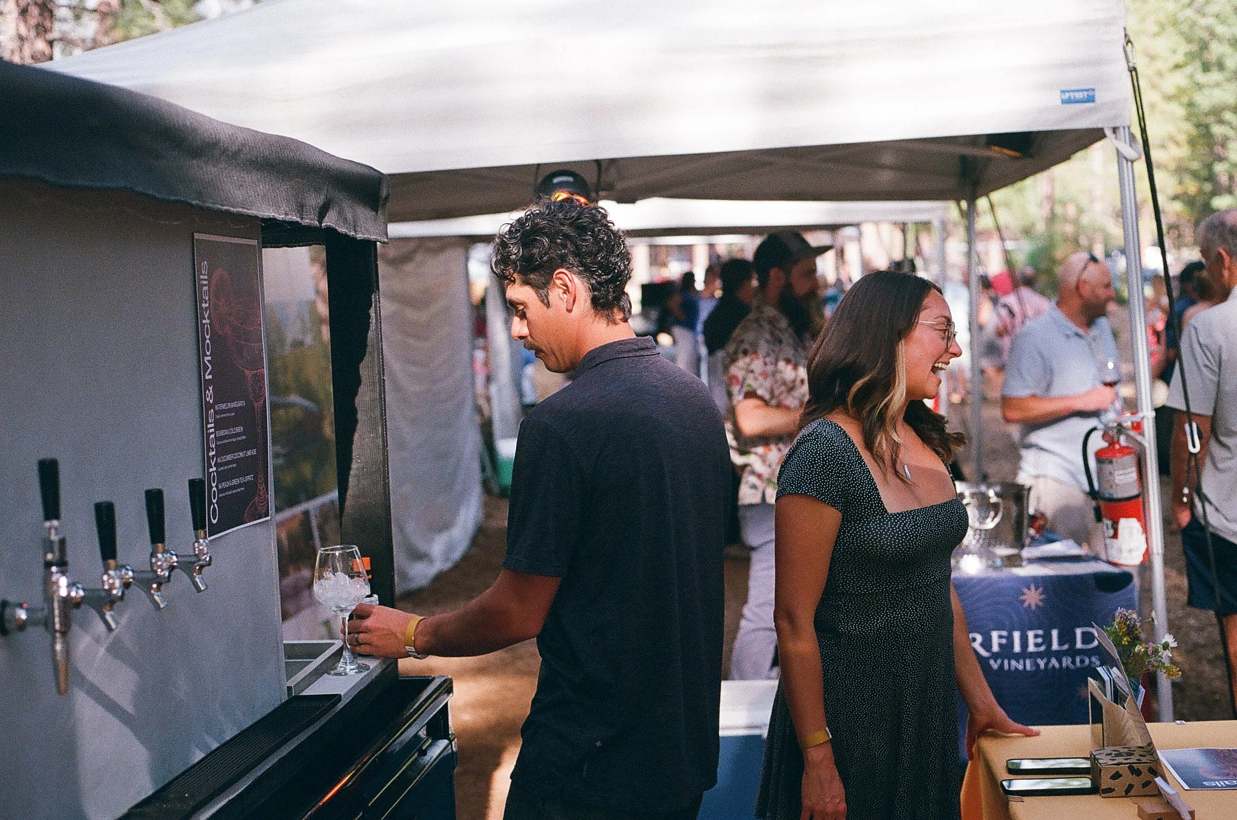 People at an outdoor event, with a man pouring wine from a tap, and a woman smiling nearby. Tents and other attendees are visible in the background.