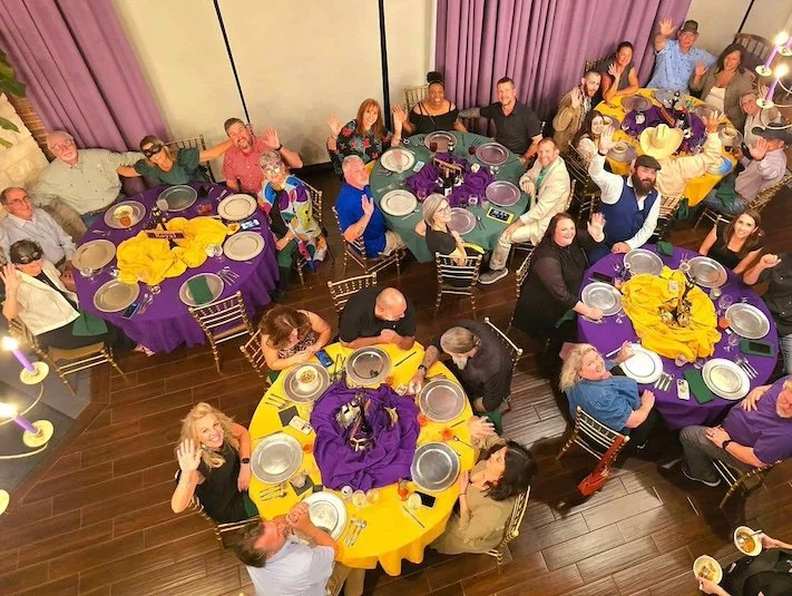 Top-down view of a diverse group of people celebrating at a party, sitting around round tables with colorful tablecloths and centerpieces, smiling and waving at the camera.