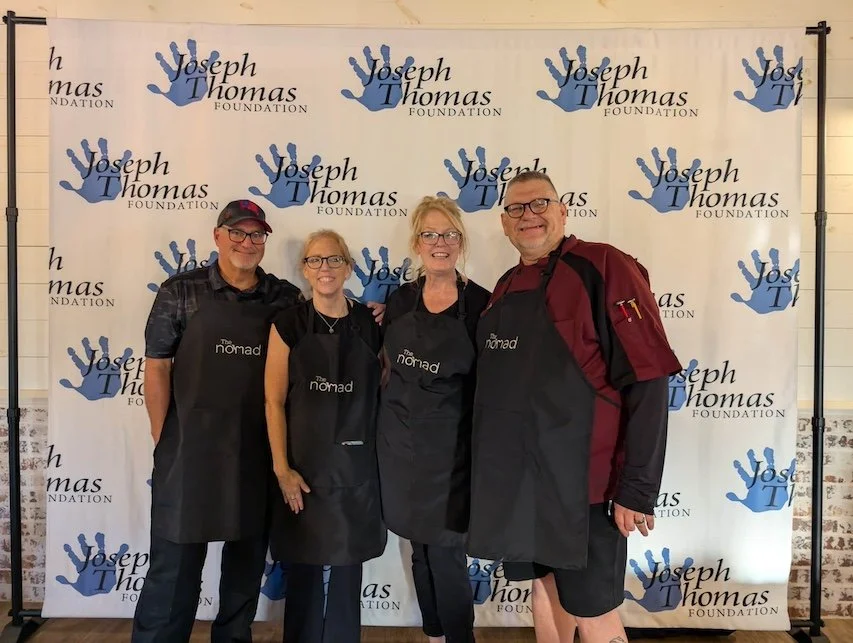Four people standing together in front of a backdrop with the Joseph Thomas Foundation logo, wearing black aprons that say "The Nomad." They are smiling and appear to be at an event or gathering.