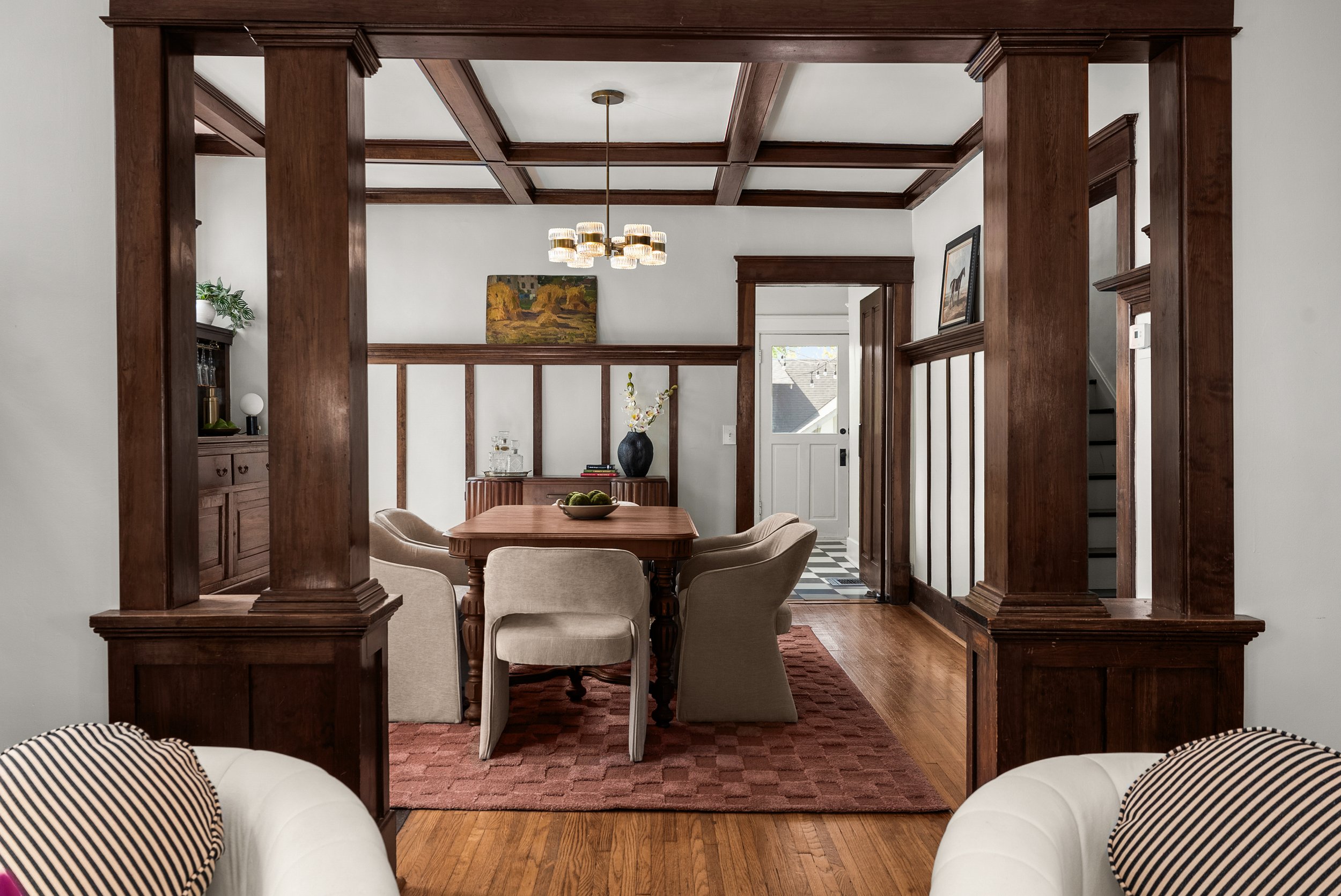 A dining room viewed through decorative wooden archways with a wooden table surrounded by six beige upholstered chairs, a red patterned rug underneath, and a wooden sideboard with a vase of flowers. The room has a white door, artwork, and a modern chandelier with multiple bulbs.