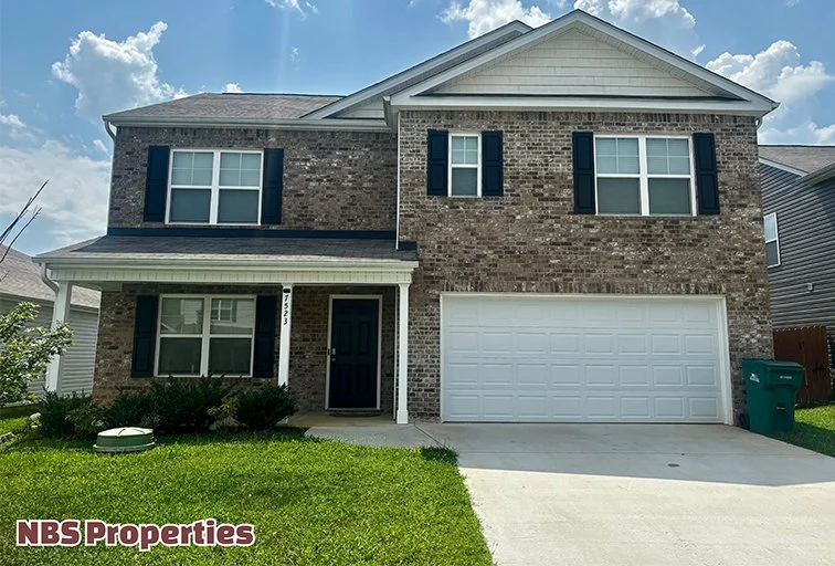 Two-story brick house with a dark front door, white garage door, and black shutters. Green lawn and a clear sky.