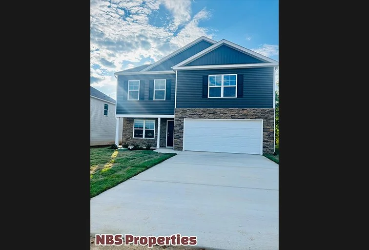 New two-story house with a dark blue exterior, stone accents, white garage door, and a concrete driveway under a partly cloudy sky.