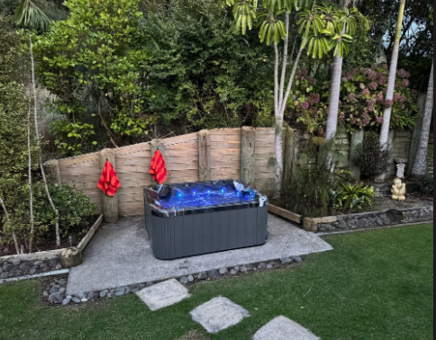 A backyard with a hot tub, palm trees, and green grass, featuring a small statue.