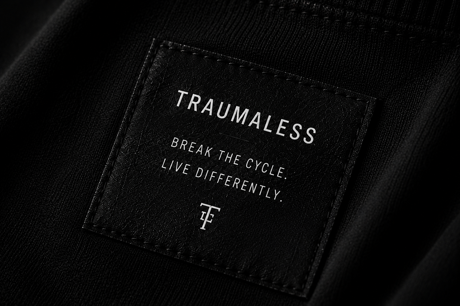 Close-up of a black leather label with white text that reads "TRAUMALess," followed by the phrases "Break the cycle. Live differently," and a small logo with the letters "TF".