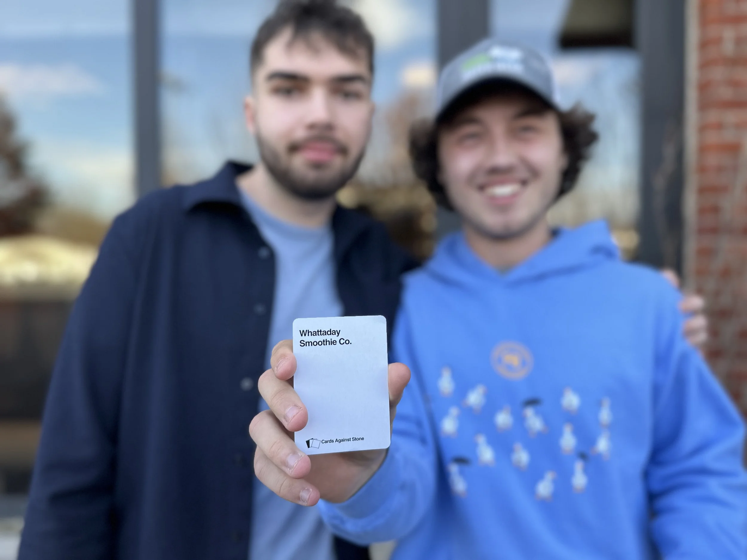 Two young men standing outdoors, smiling, with one holding a small card that reads 'Whattaday Smoothie Co.'