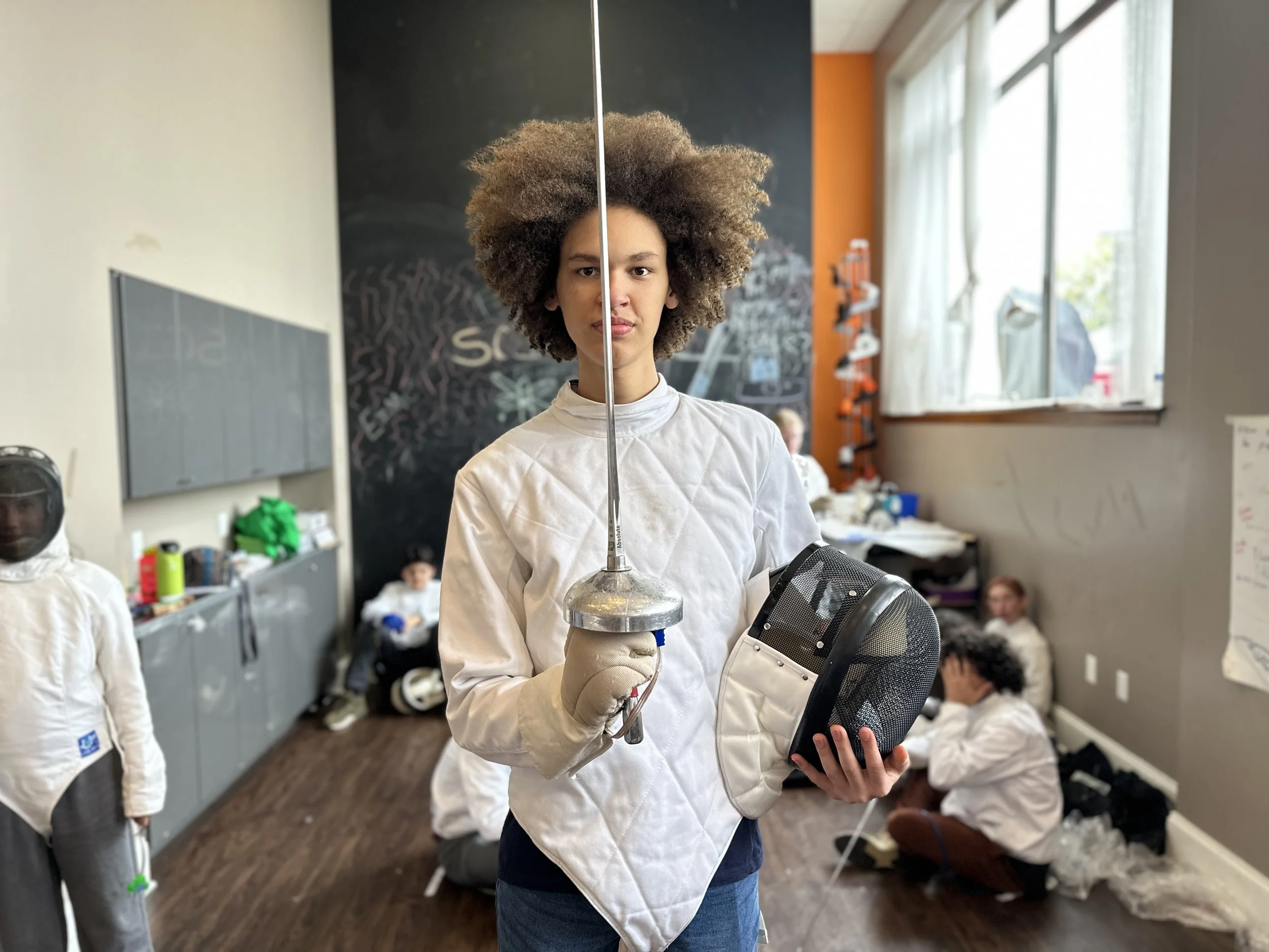 Young woman standing in a classroom holding a fencing mask and a foil, with other children in fencing gear in the background, some sitting on the floor and one wearing a helmet, girl with glasses sitting near the window.