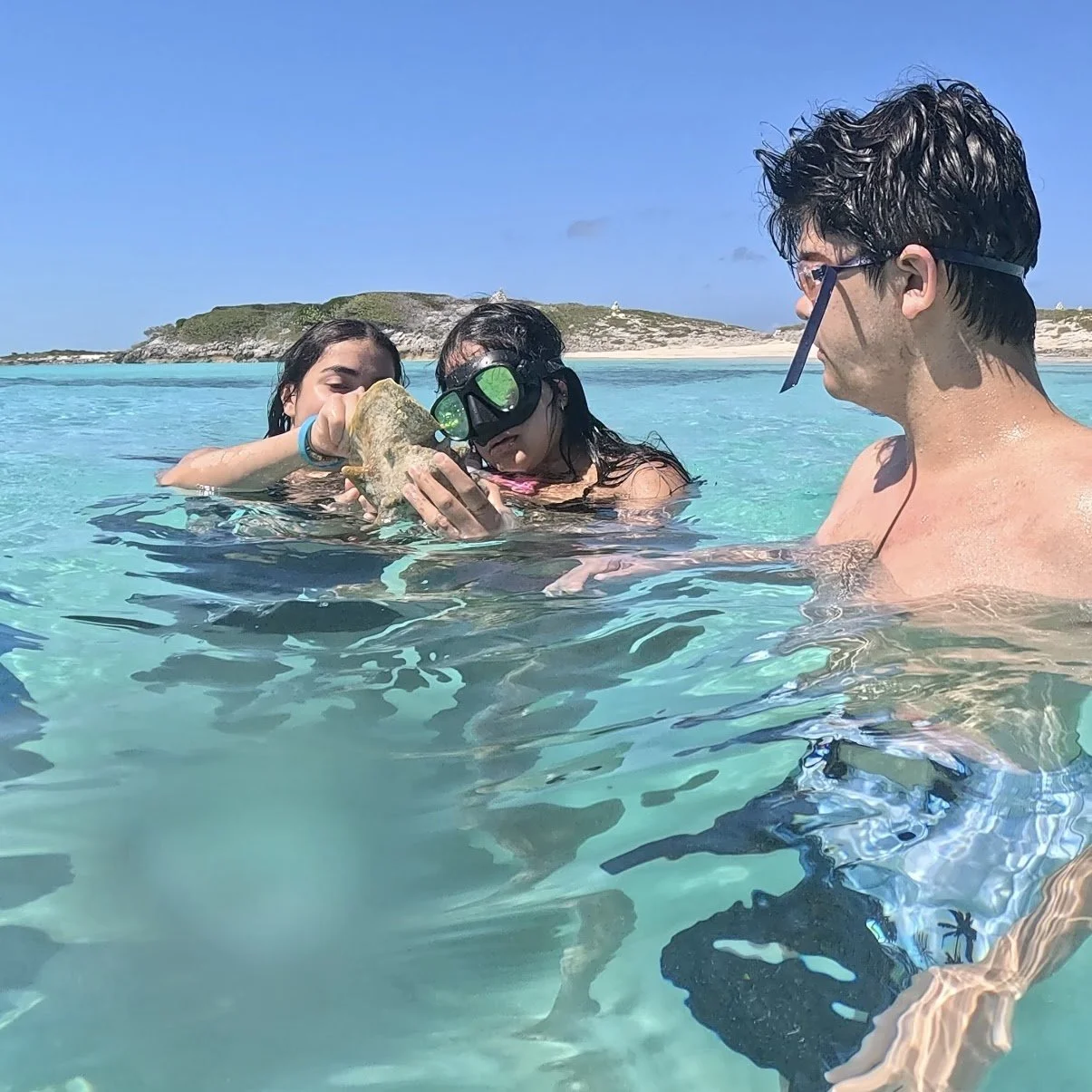 Man holding a small crab with blue claws out towards camera, smiling, outdoors, with a blurred beach background.