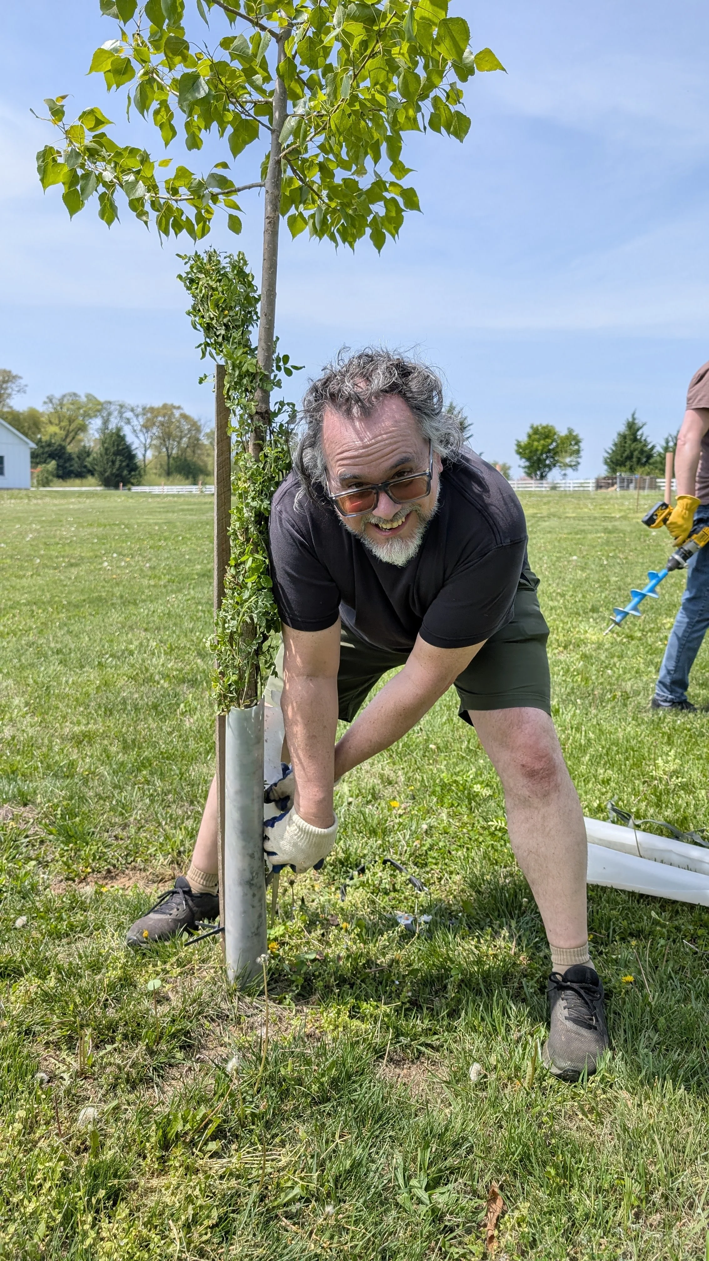 Two women smiling and taking a selfie outdoors, standing next to a large tree with lush green landscape and a stone wall in the background.