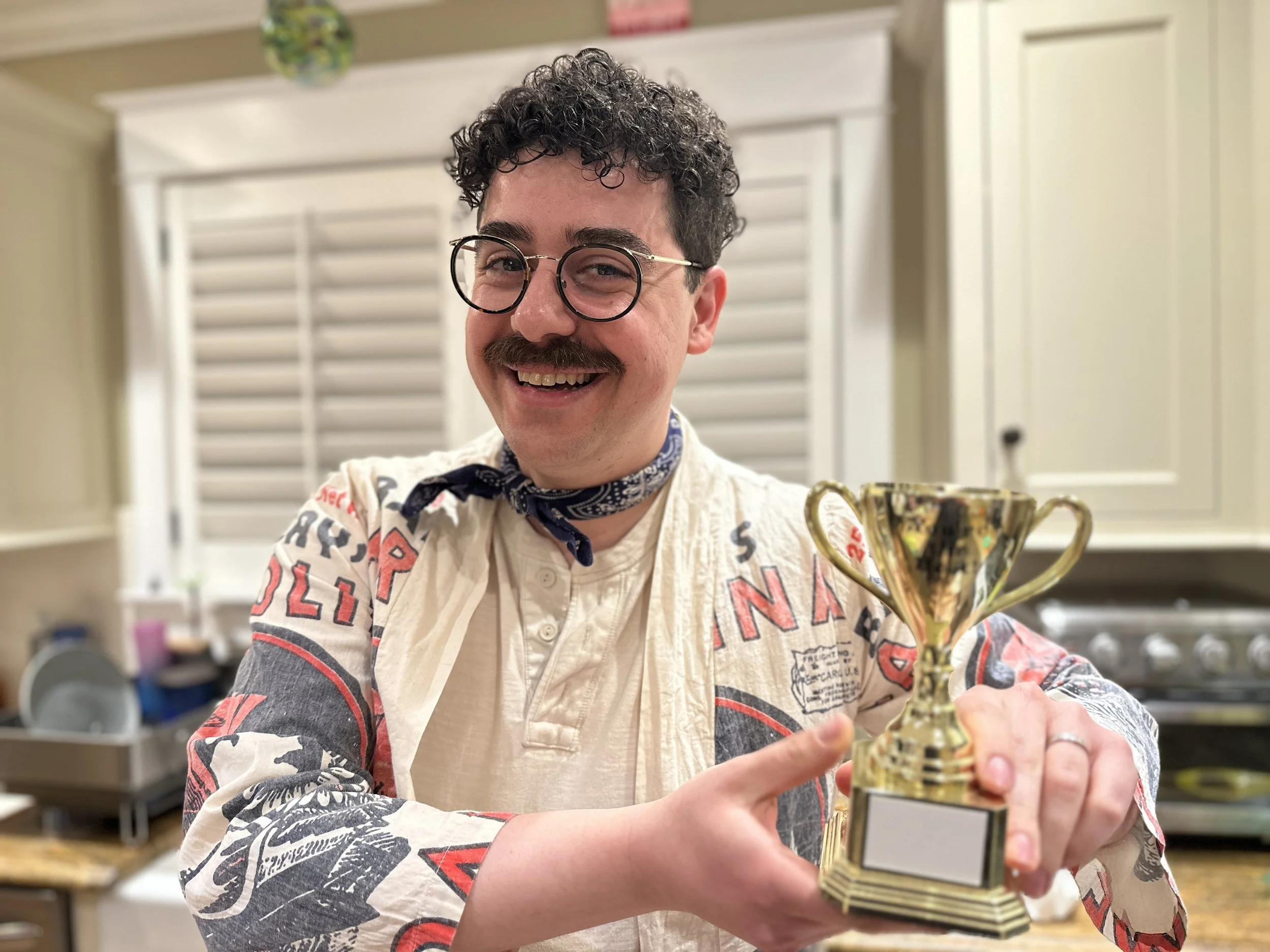 A man with glasses, curly hair, and a mustache smiles while holding a gold trophy in a kitchen.