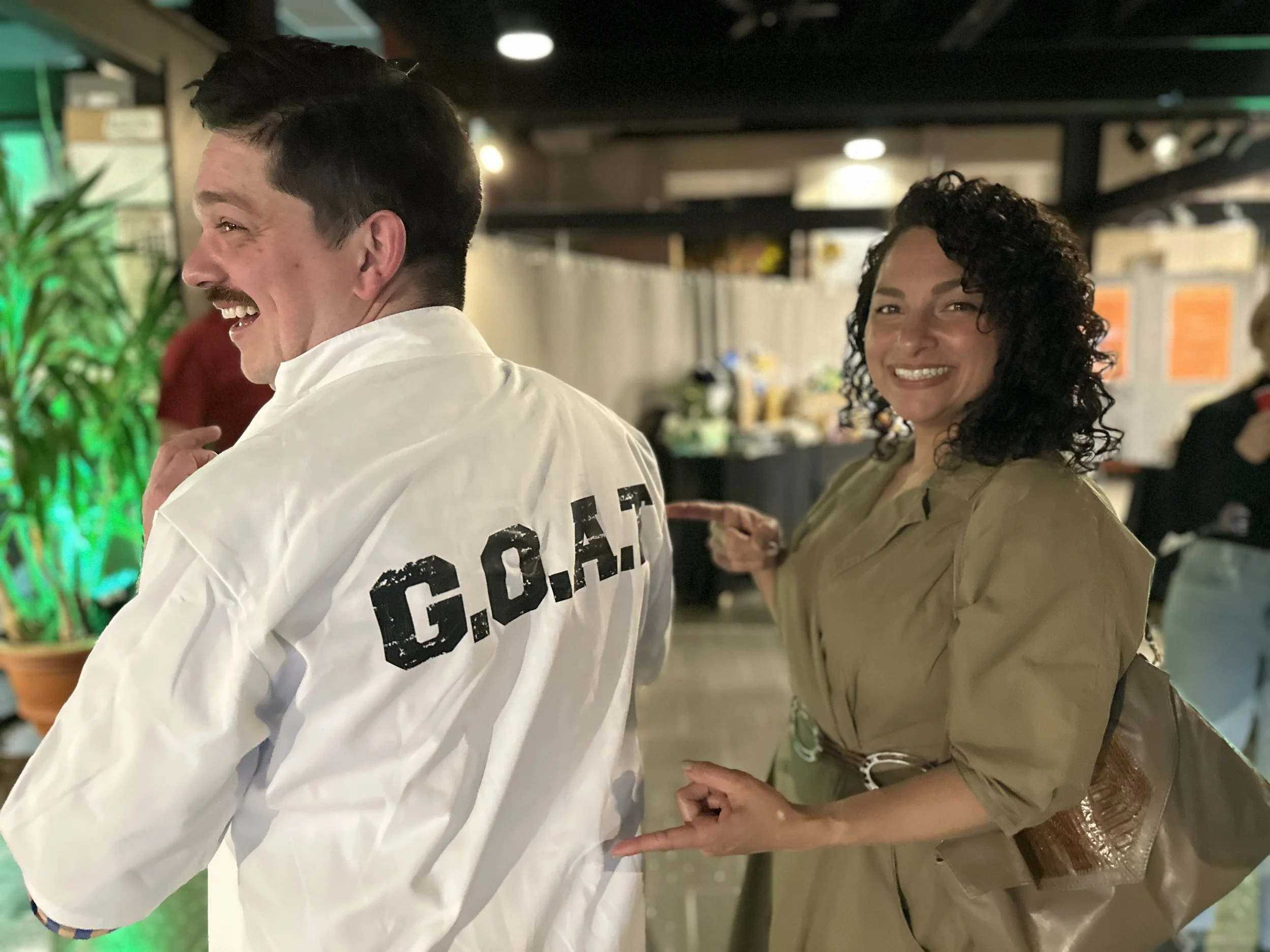 A man wearing a white jacket with 'G.O.A.T' printed on the back and a woman in a tan dress or coat smiling and pointing at him at an indoor event with blurred background and people.