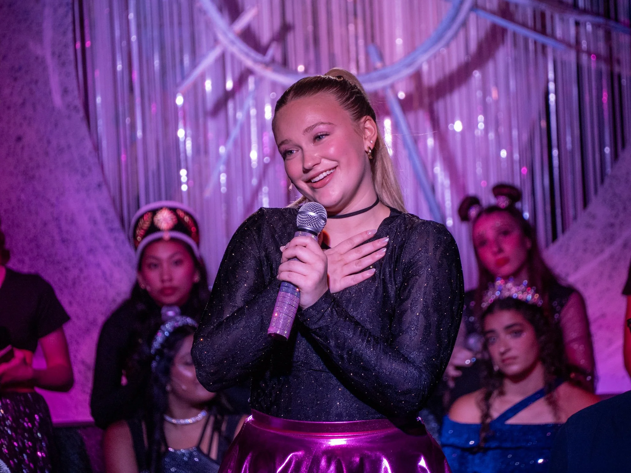 A man with curly black hair, a mustache, and glasses smiling outdoors, holding a green mug and a vinyl record album titled "Jascha Heifetz Favorite Encores" featuring a picture of a violin on the cover. He is wearing a brown jacket, green pants, a striped shirt, and a patterned scarf around his neck, standing in front of a red brick building with parked cars.