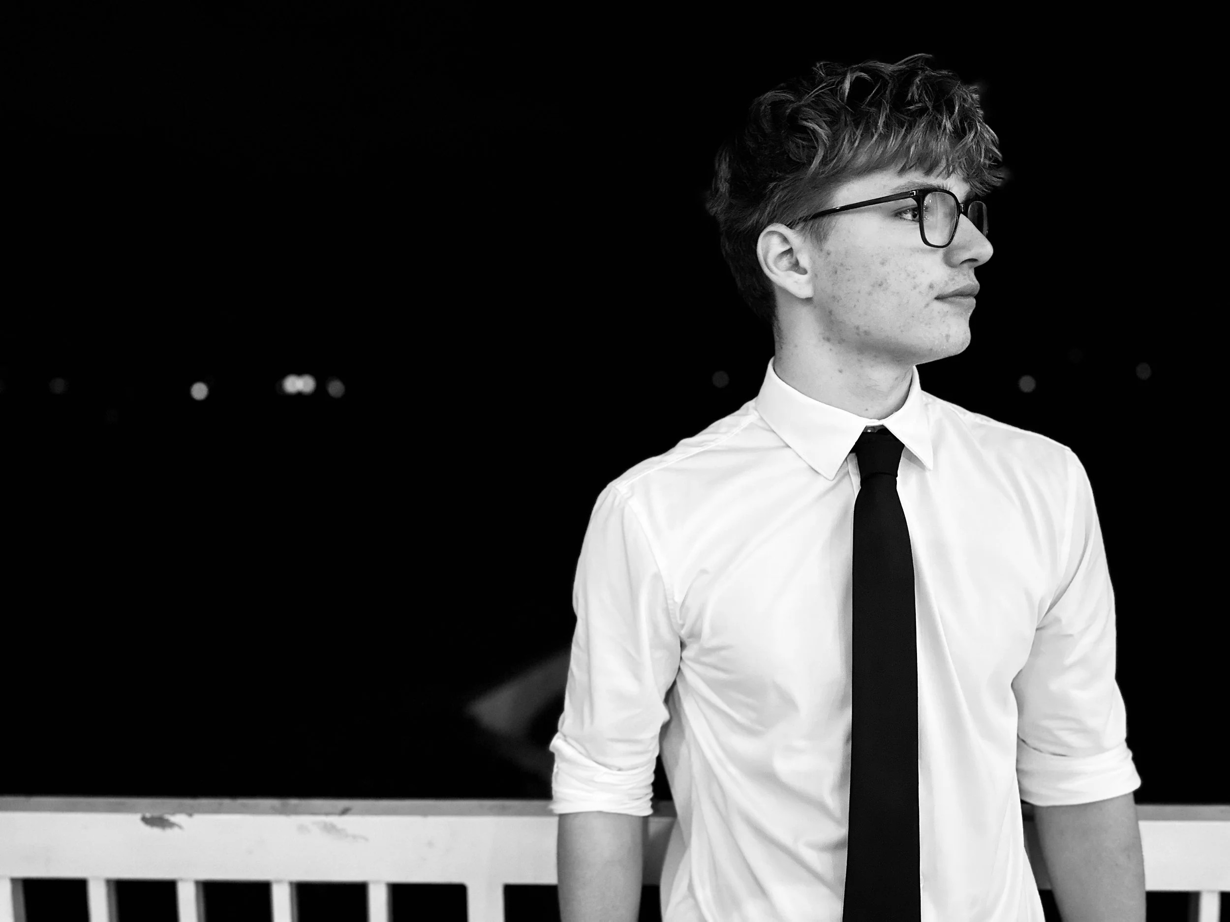 Young man with glasses in a white shirt and black tie standing outdoors at night, looking to the right, with a dark background and faint lights in the distance.