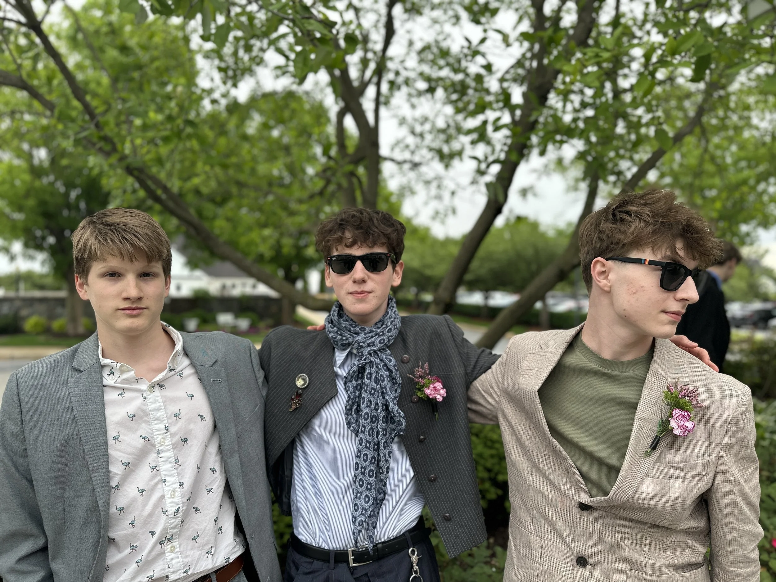 Three young men standing outdoors with green trees in the background, wearing suits and sunglasses, with the middle one having a scarf and boutonniere.